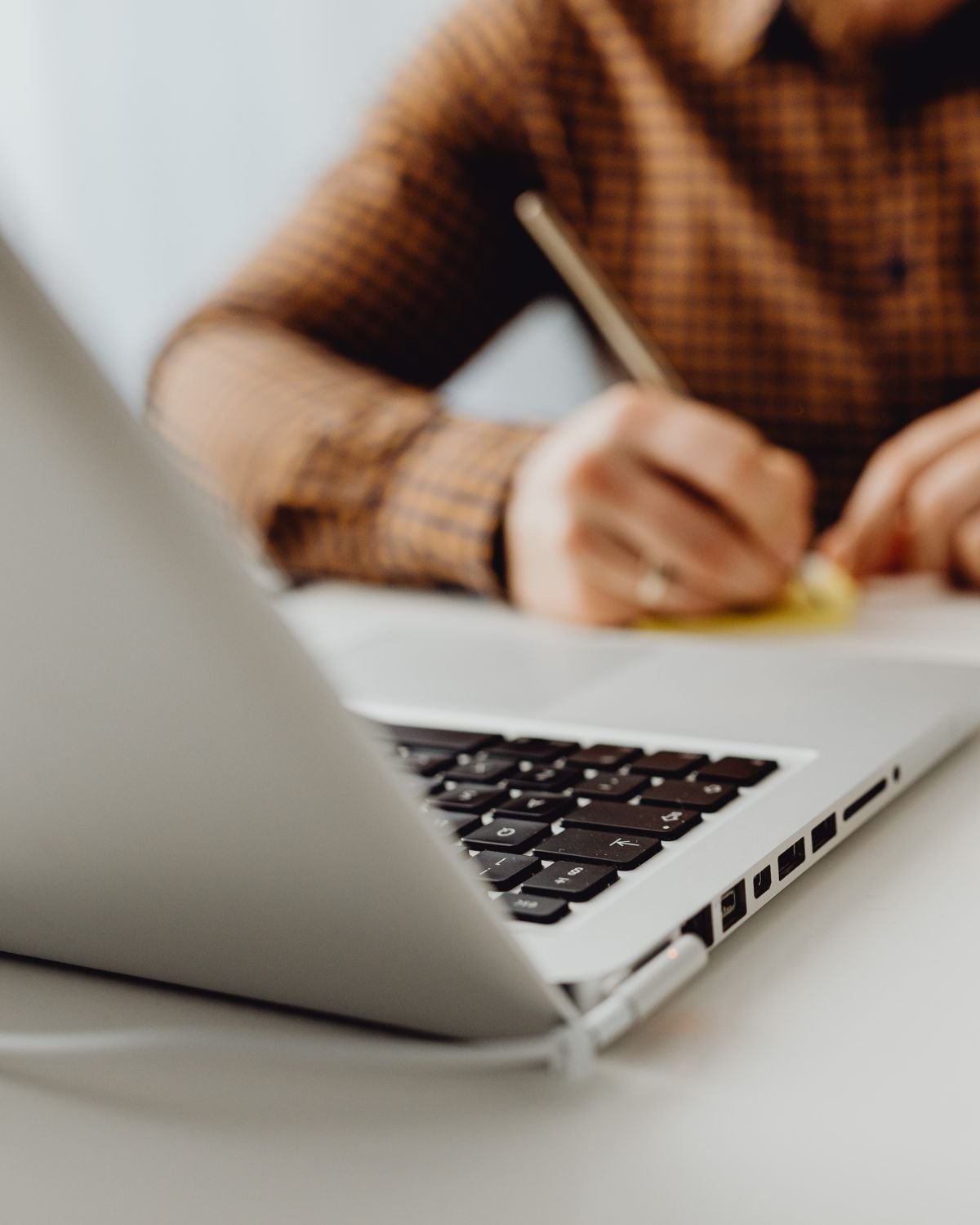 Person writing notes on paper with a pen, laptop on desk in foreground.