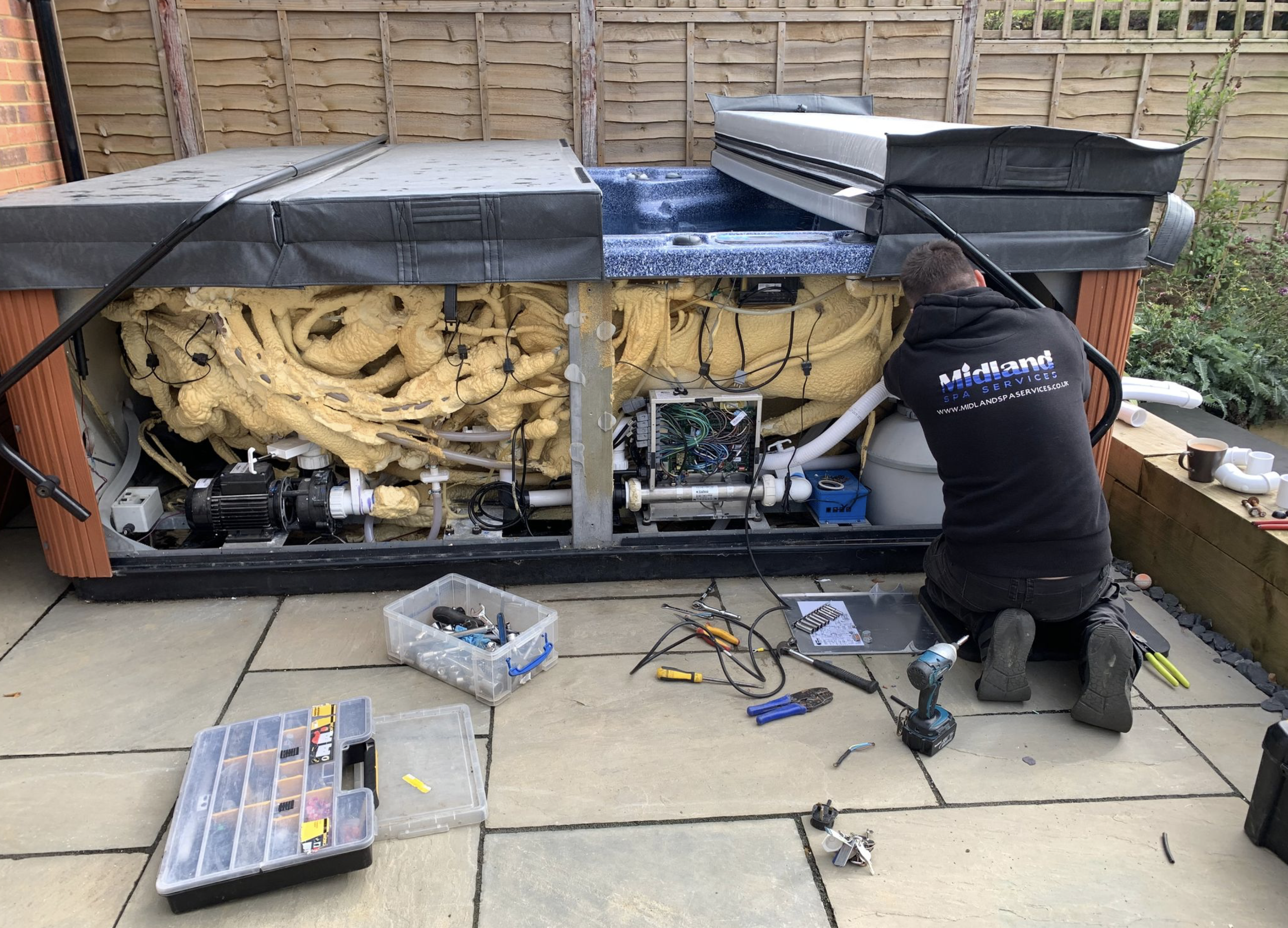 A technician installing or repairing a hot tub or spa outdoors on a patio, with tools and equipment around him.