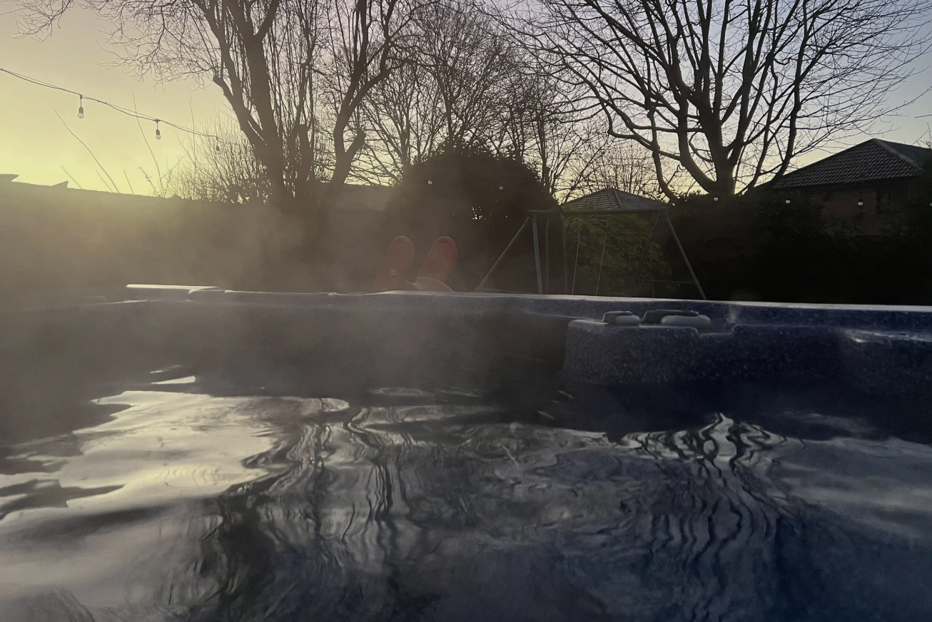 View from inside a hot tub with steam rising, showing leafless trees and houses in the background during sunset, with a person's hand making a peace sign.