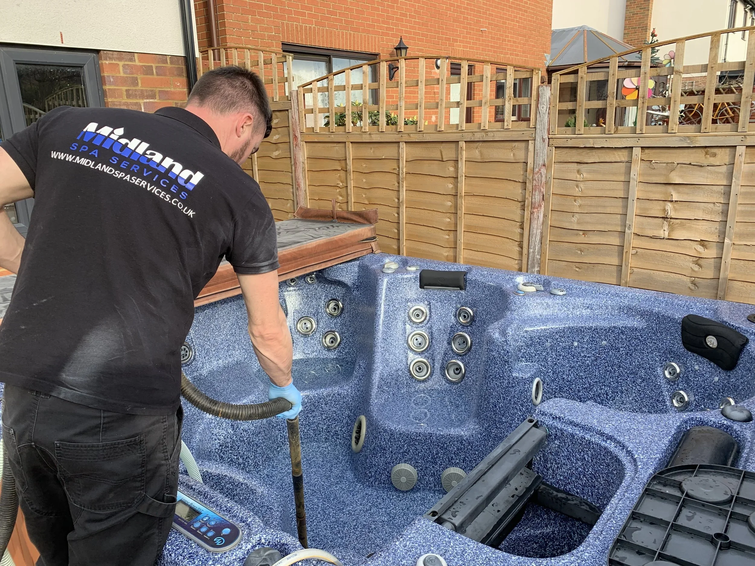A technician cleaning a blue hot tub outdoors on a patio with a wooden fence and brick house in the background.