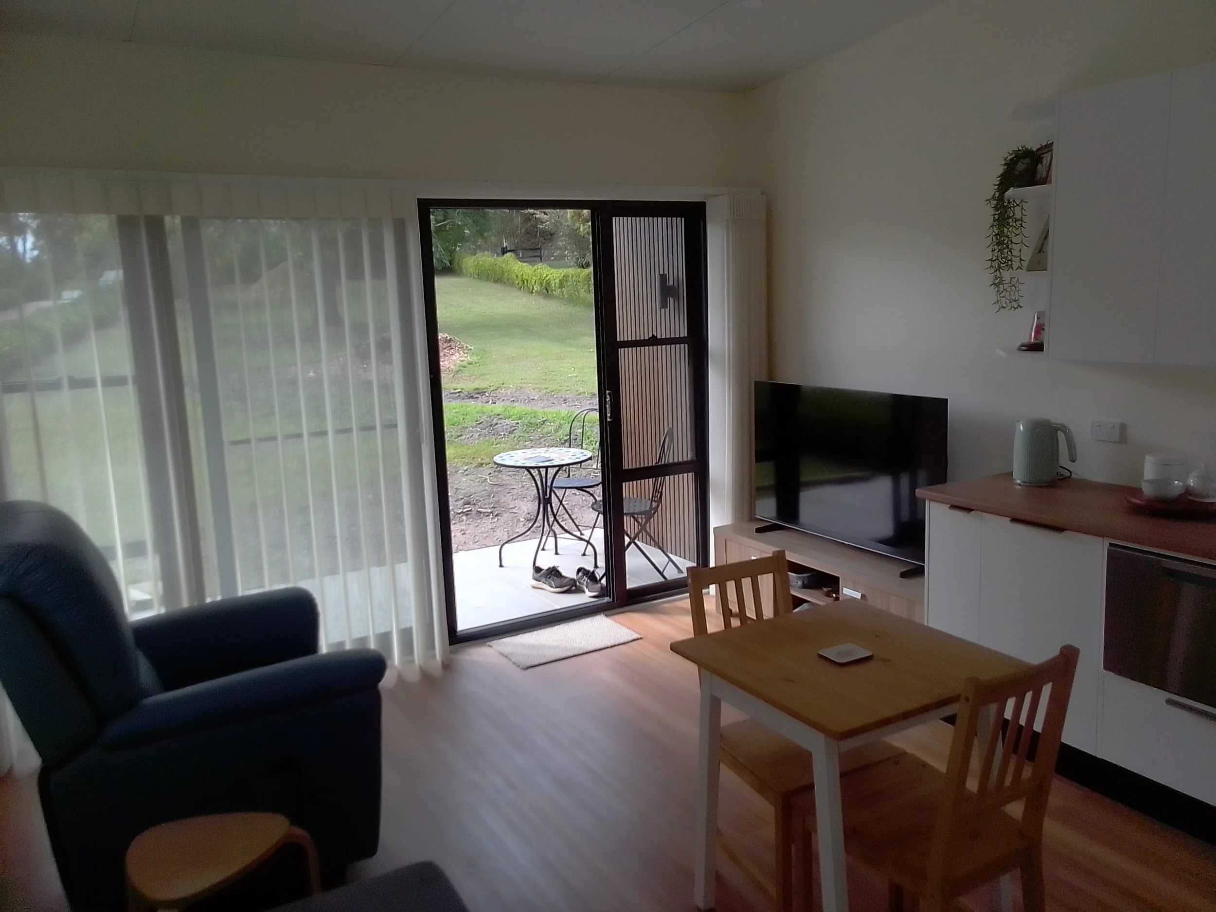 Interior of a living room with a sliding glass door leading outside, a black armchair, a small table, a television on a stand, and white cabinets with plants and books on the wall.