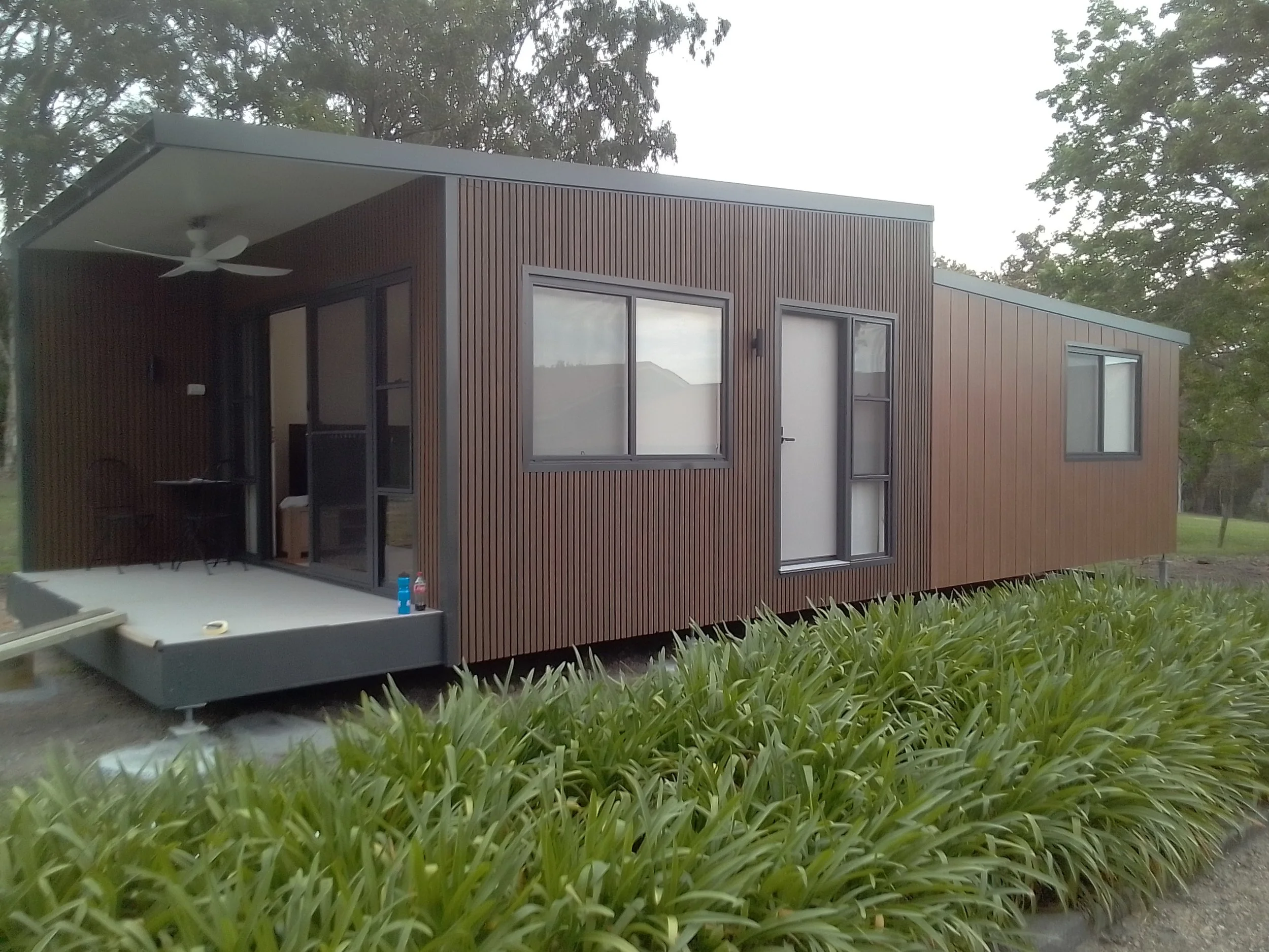 Modern tiny house with wooden exterior, glass doors, and large windows, surrounded by greenery and trees.
