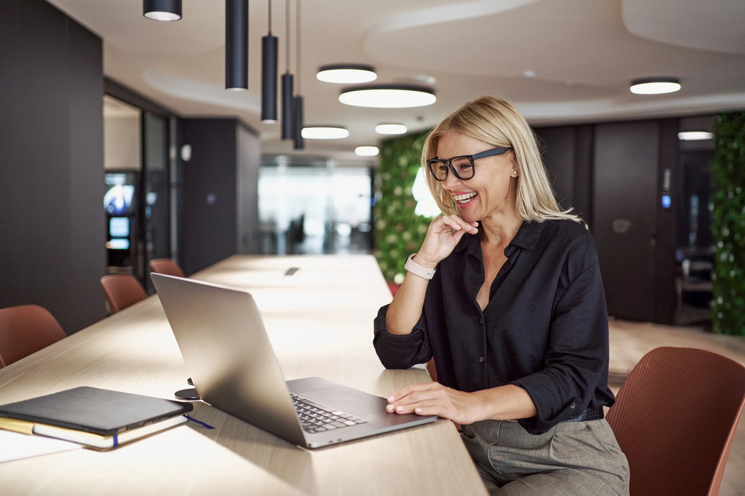 Mulher sorridente com cabelo loiro e óculos escuros usando roupas pretas e cinzas, sentado em uma mesa de reunião com laptop, caderno e caneta em ambiente de escritório moderno.