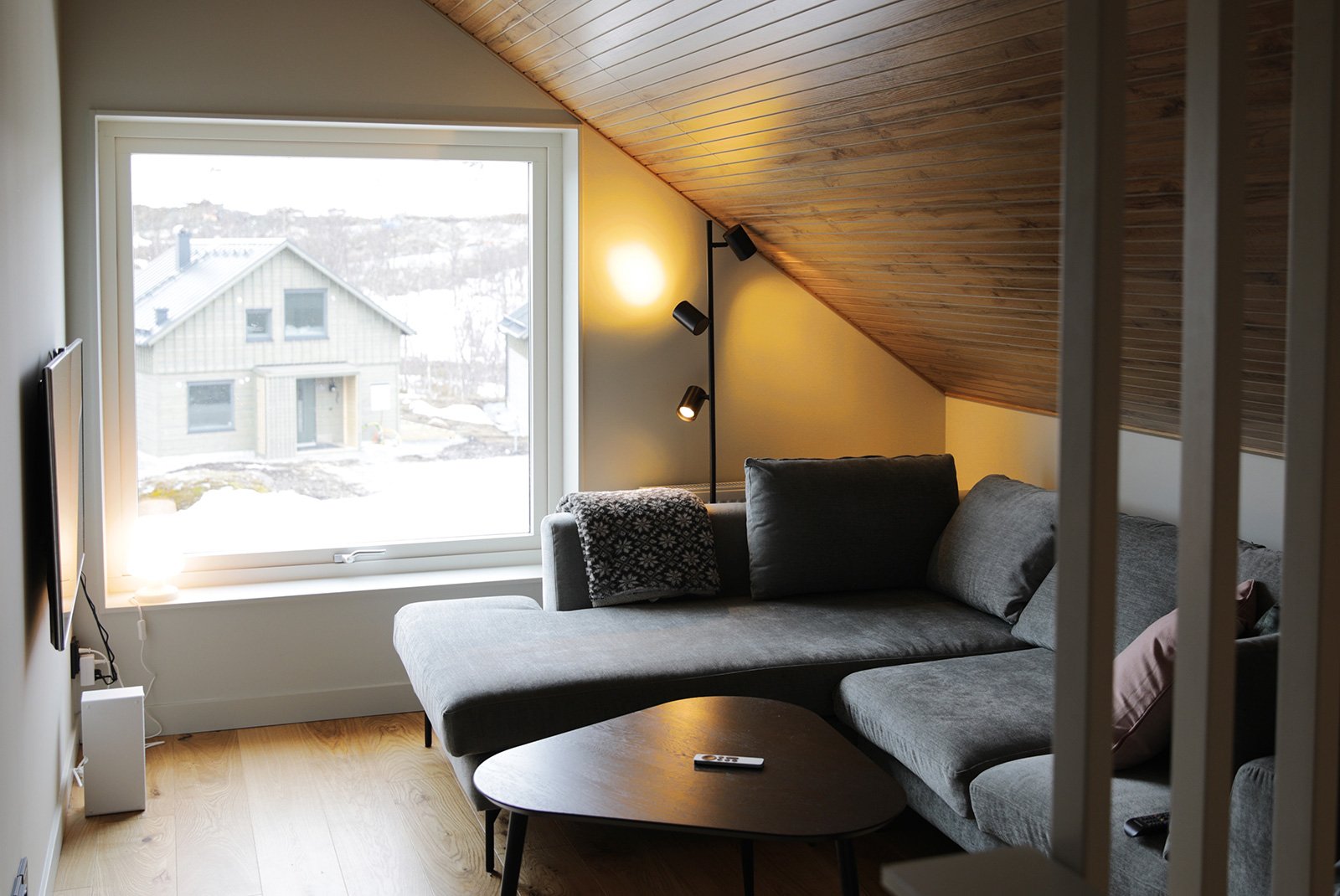 Vardagsrum med grå soffa, träfönster, slät vägg och trämålning i taket.