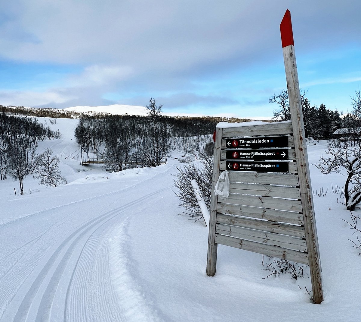 Snösnöig skidspår och en skylt i vinterlandskap med träd och kullar i bakgrunden.