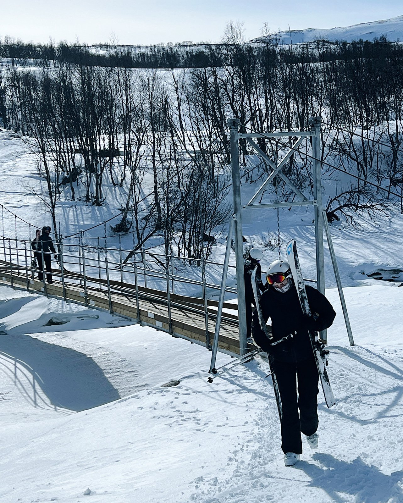 Person i skidutrustning bär skidor och håller en andra skidpar, går mot en snöig bro med andra skidåkare längre bort, i ett vinterlandskap med snöklädda träd och berg i bakgrunden.