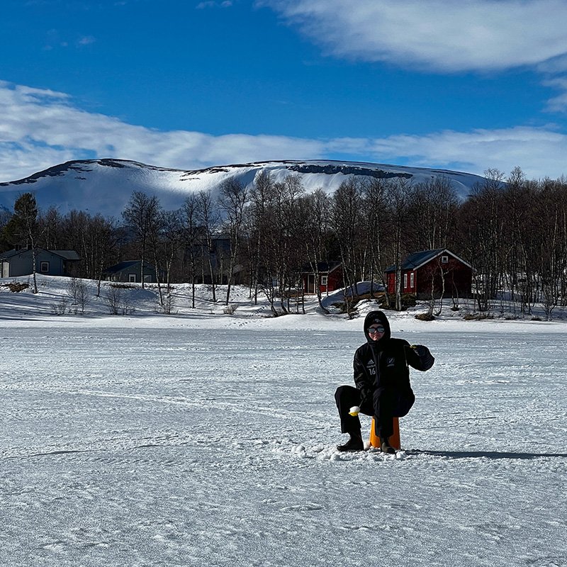 Person i vinterkläder sitter på en stol på snötäckta landskapet med snötäckta berg och vilande trähus i bakgrunden, under en blå himmel med moln.