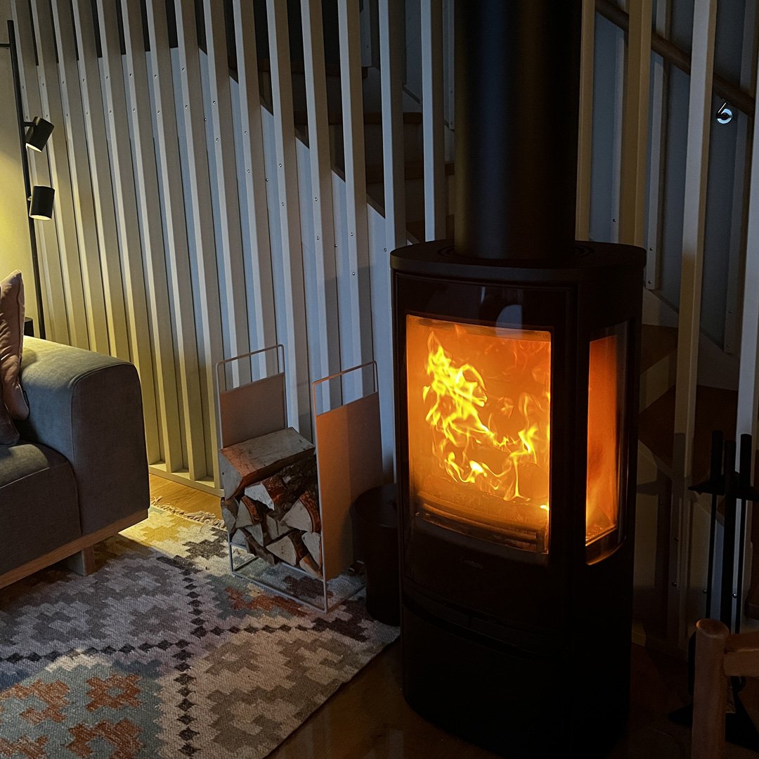 En modern eldstad med öppet fönster som visar en brinnande eld, placerad i ett vardagsrum med en soffa, matta och en liten pappersbrasa