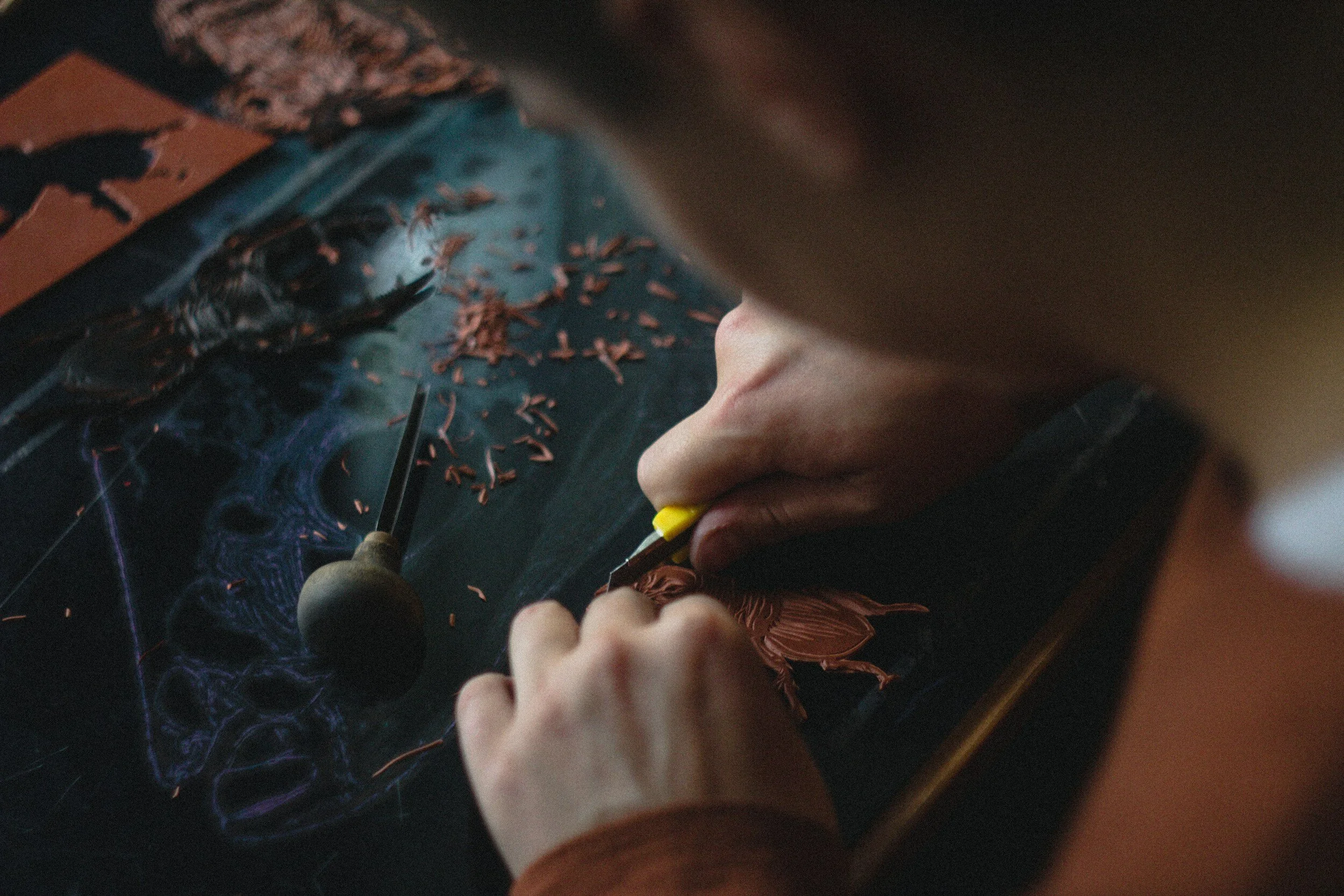 Close-up of a person carving a design into a block of linoleum using a carving tool, surrounded by shavings.