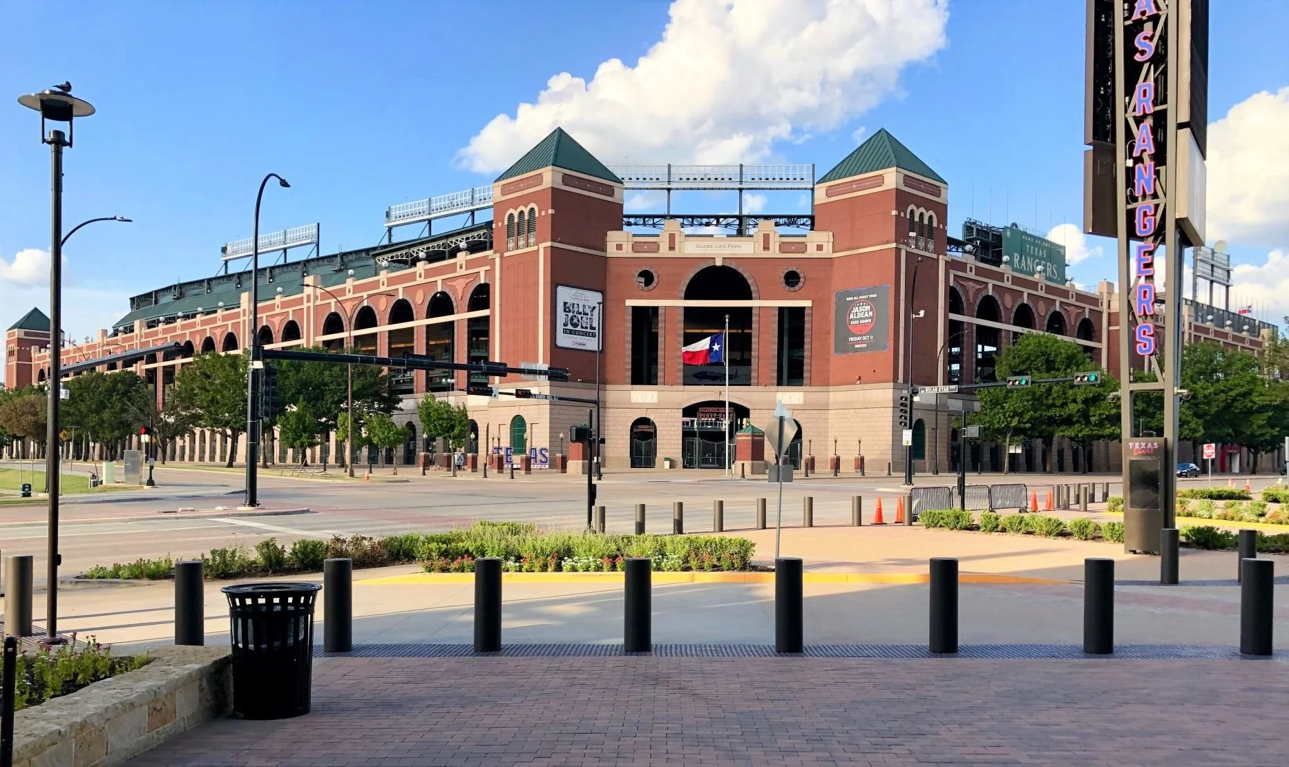 A large stadium with red brick exterior, green roofs, and arches, located in an open area with streets, trees, and a flag in front, under a partly cloudy sky.