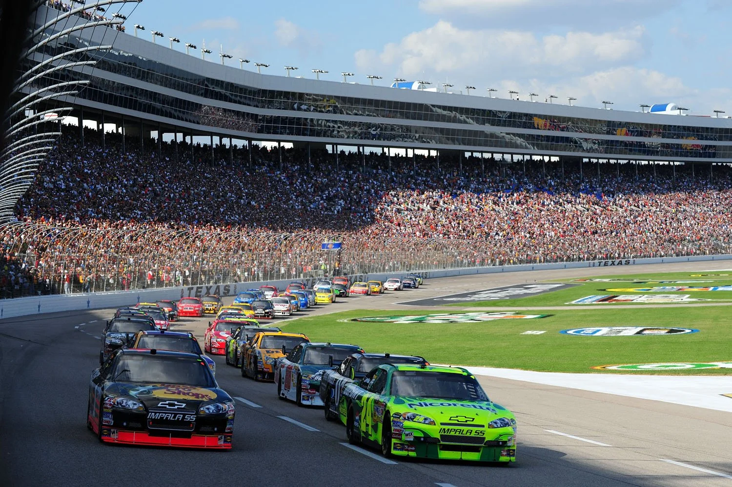Race cars on a track at Texas Motor Speedway during a daytime race with spectators in the grandstand.