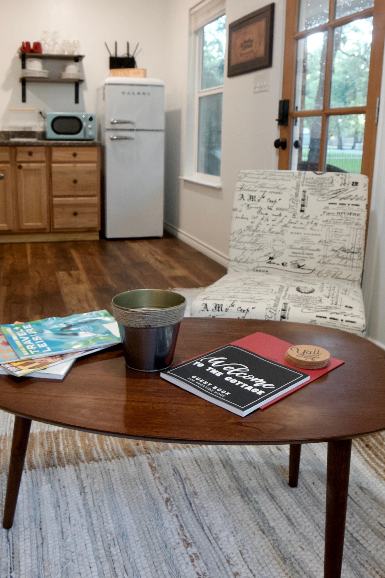 A cozy living room corner with a wooden coffee table holding magazines, a black cup with a green interior, a guest book titled 'Welcome to the Cottage,' and a wooden coaster. In the background, there is a beige armchair with script-patterned upholstery, a window, and a door leading outside, with a kitchen area featuring a vintage-style refrigerator, a microwave, and open shelves with dishes.