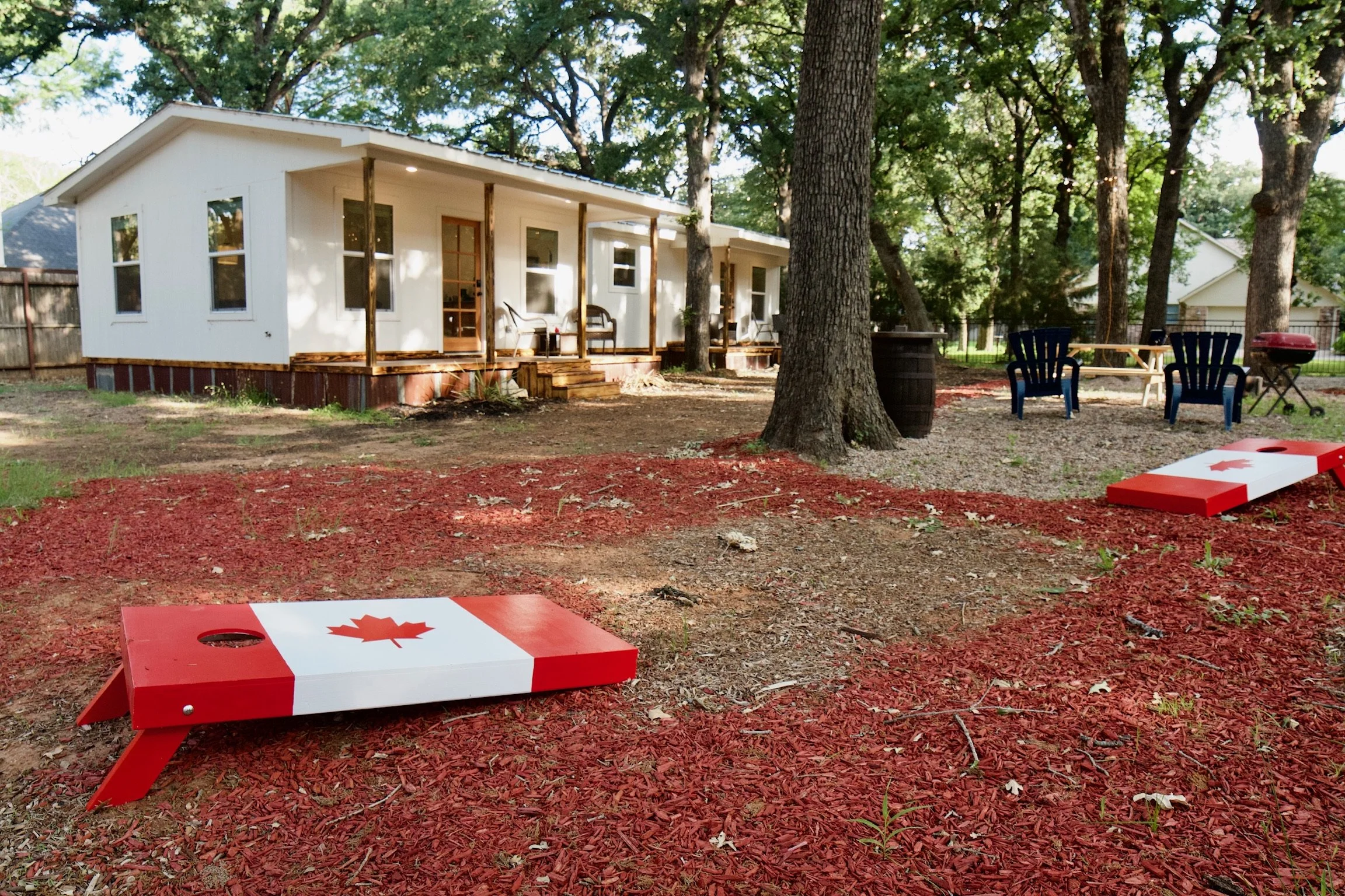 Backyard with a white house, trees, seating area, and two cornhole game boards decorated like Canadian flags.
