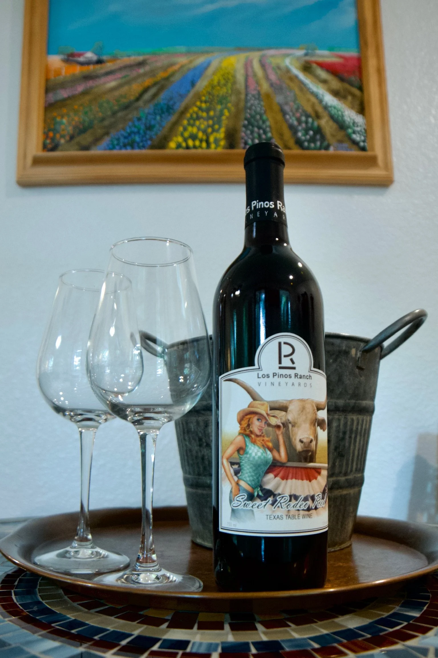 A bottle of Los Pinos Ranch Vineyards Sweet Rodeo Red wine, three empty wine glasses, and a metal ice bucket on a round wooden tray, with a colorful pieced mosaic border, in front of a wall with a framed painting of a field of tulips.