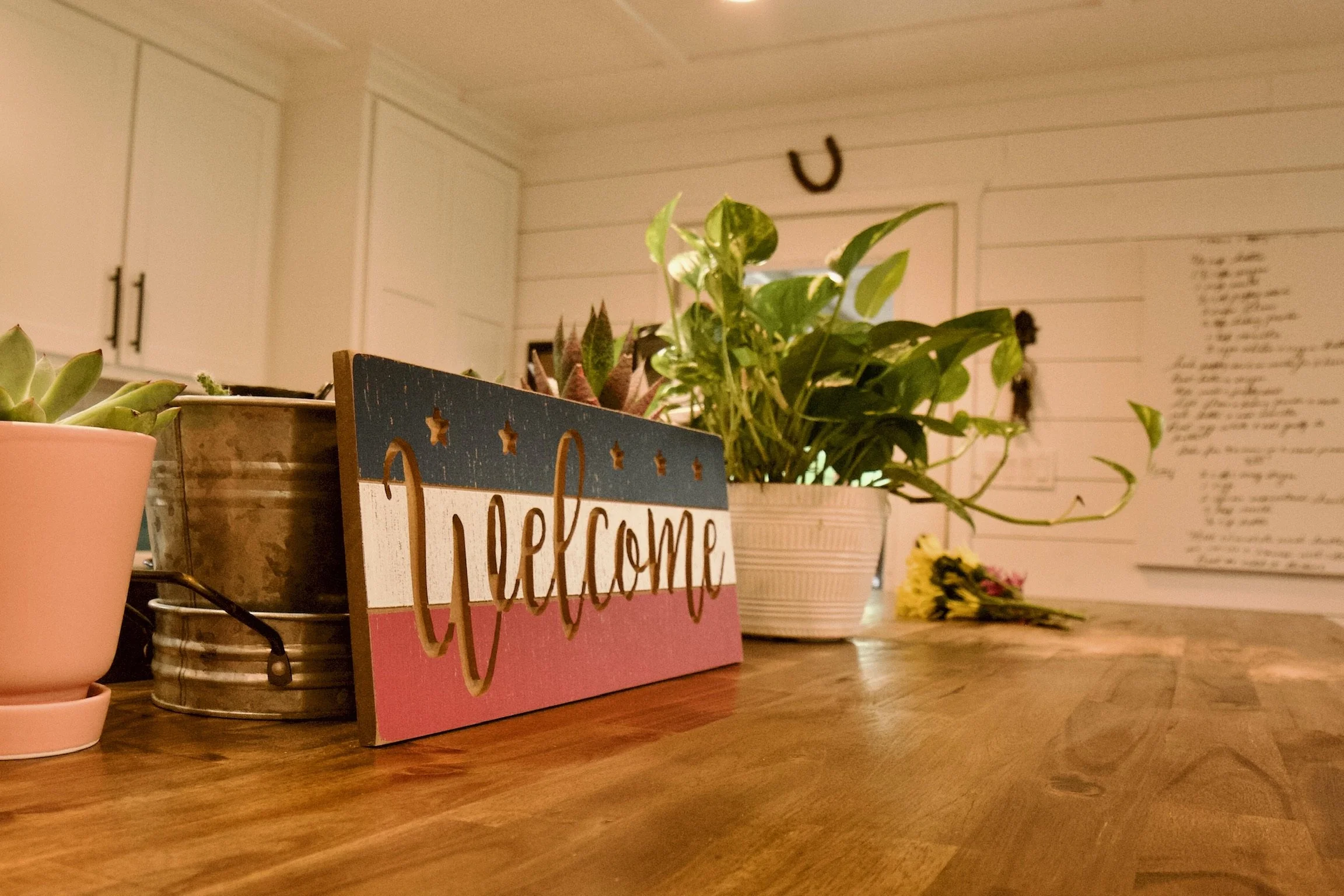 Wooden table with plants, a decorative 'welcome' sign, and flowers in a cozy, white-washed room.