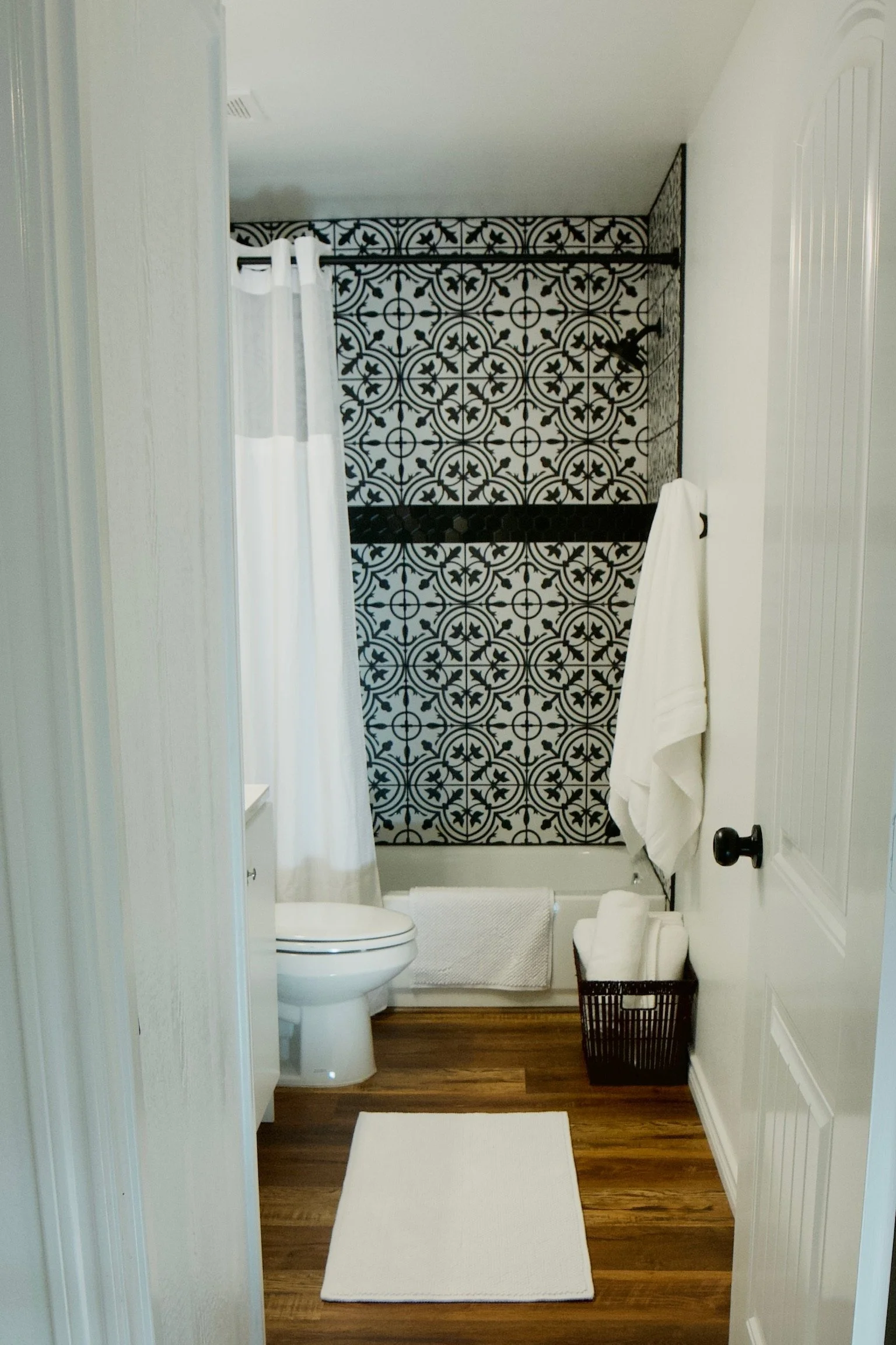 View of a bathroom with a white toilet, black and white patterned shower wall, white curtain, white towels, a white bath mat, and a wicker basket on a wooden floor.