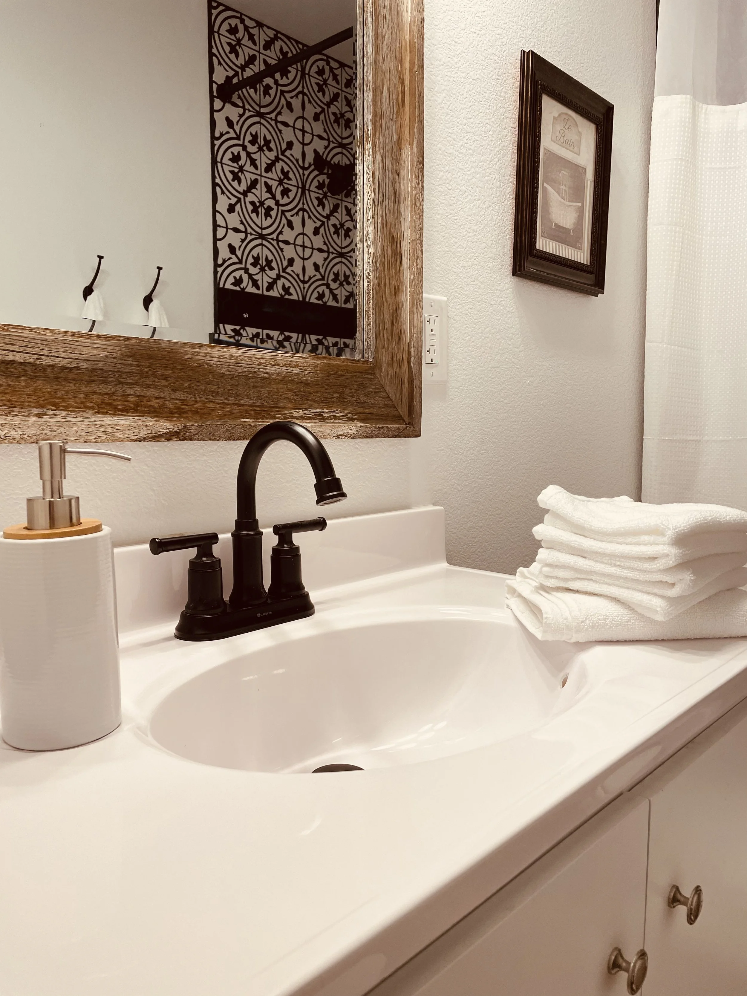 Bathroom sink with a black faucet, a stack of white towels on the right, a white soap dispenser on the left, and a wooden-framed mirror above the sink. There is a framed picture on the wall and a towel hanging nearby.