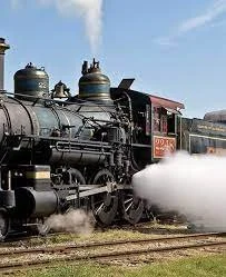 A vintage steam locomotive emitting steam as it moves along train tracks under a blue sky.