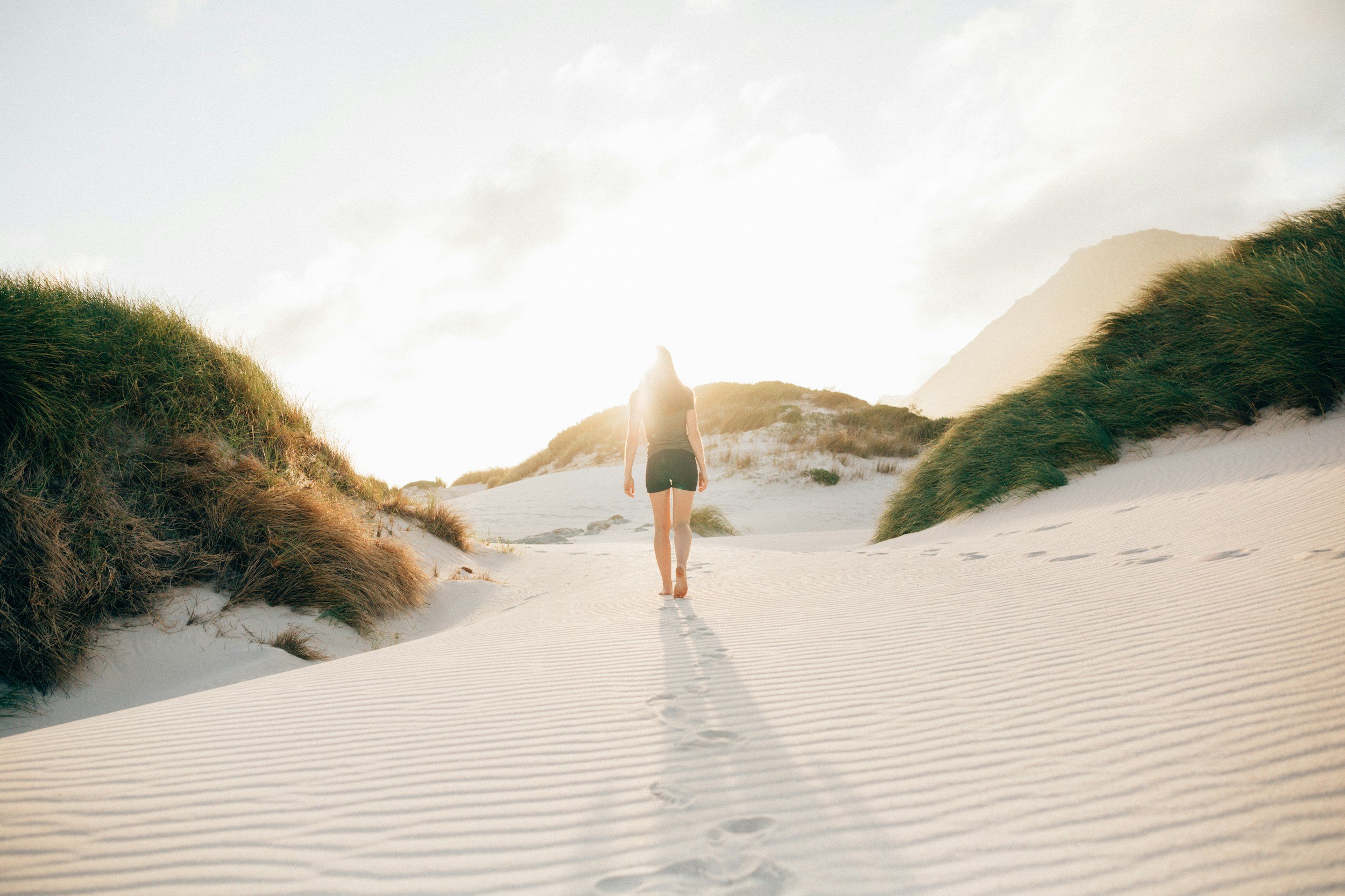 Une femme marche sur une plage de sable blanc, entourée de dunes avec de l'herbe, sous un ciel en partie nuageux, avec le soleil qui brille derrière elle.