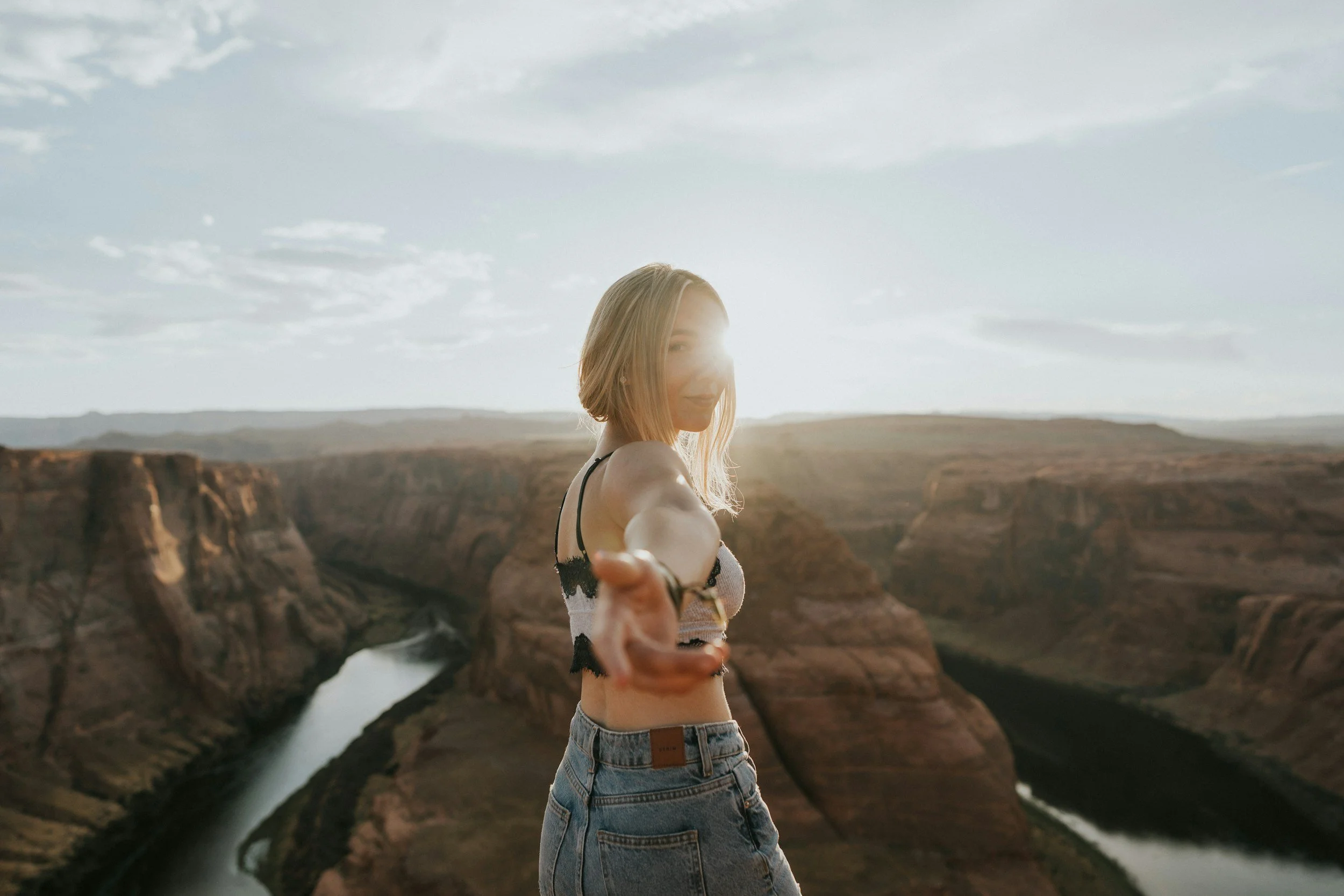 Jeune femme souriante avec une main tendue vers la caméra, paysage de canyon avec rivière au fond, soleil de coucher ou lever, vêtements décontractés
