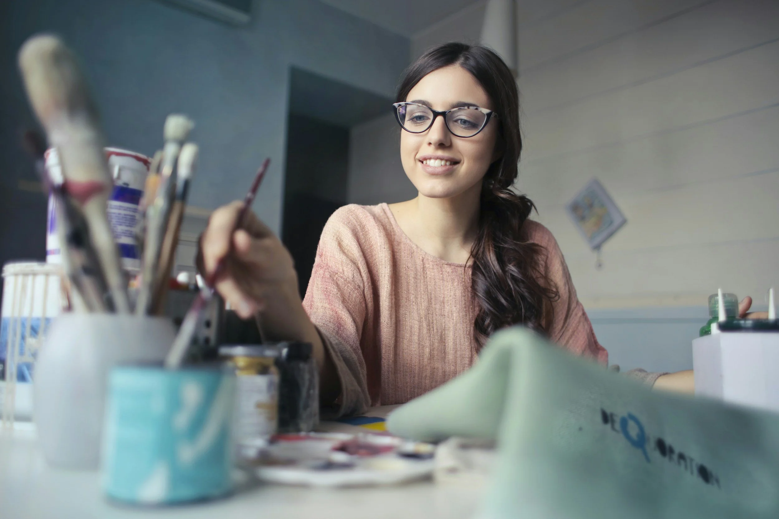 Jeune femme portant des lunettes regarde au-dessus d'une table avec des fournitures artistiques, dans une pièce lumineuse.