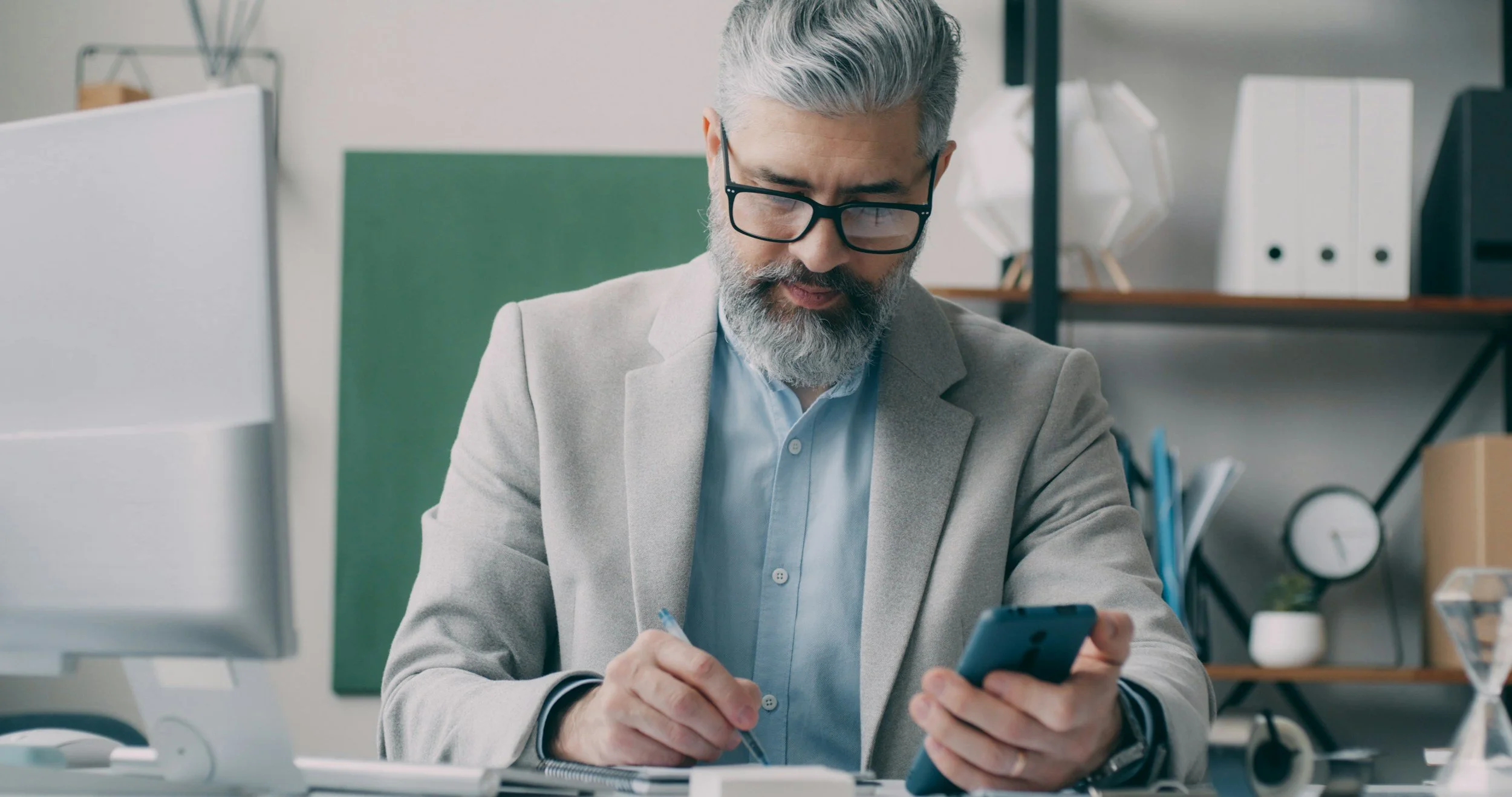 Un homme d'âge moyen avec des cheveux gris et une barbe, portant des lunettes noires, consulte son téléphone portable dans un bureau moderne. En arrière-plan, il y a des étagères avec des classeurs blancs et des décorations.