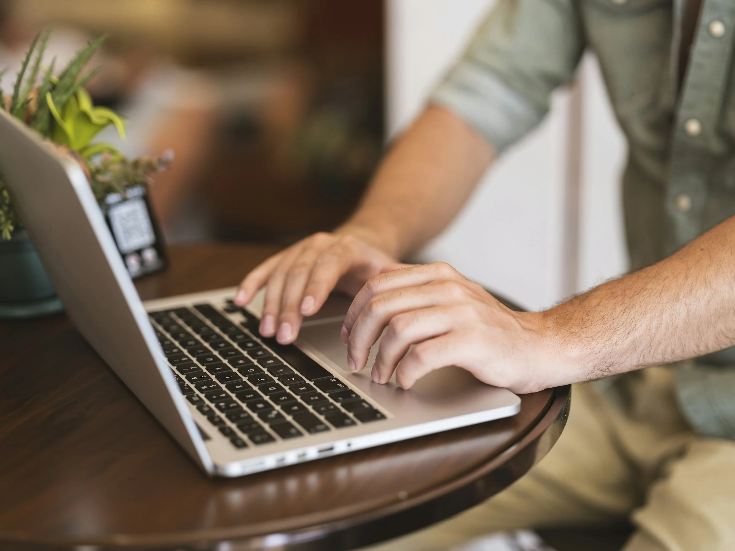 Personne tapant sur un ordinateur portable sur une table en bois, avec une plante verte à côté.