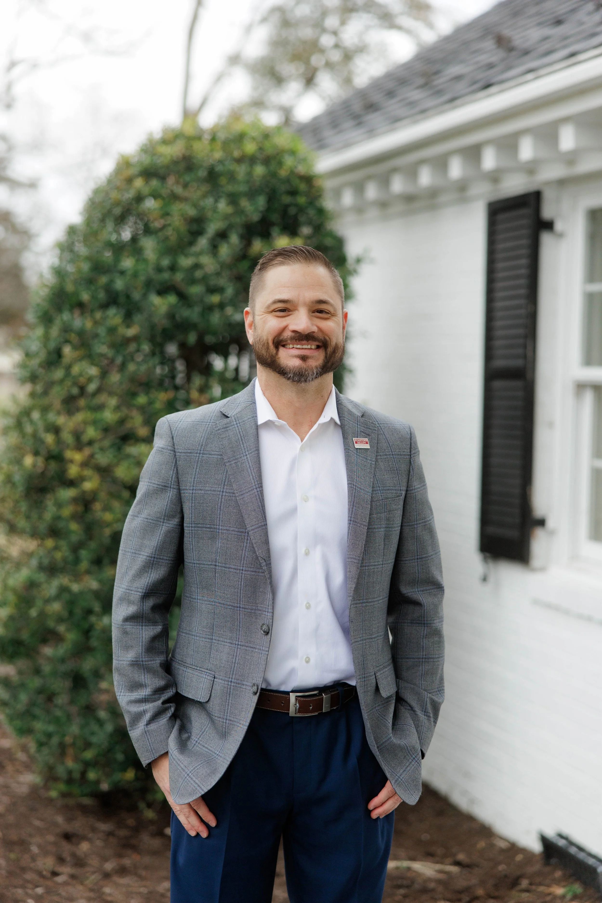 A man in a blue checkered blazer and white shirt standing outdoors with a blurred garden and greenhouse in the background.