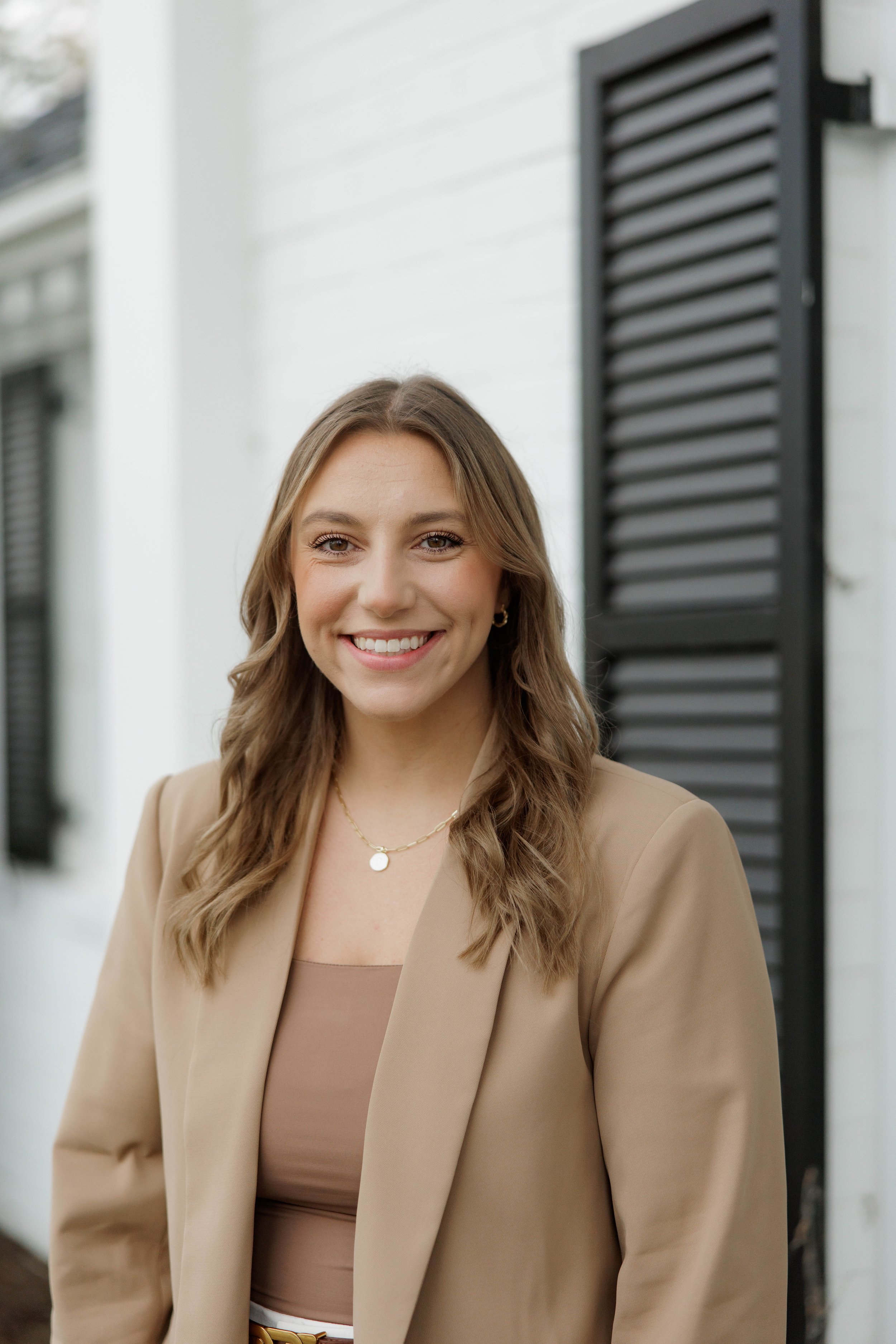 A woman with wavy light brown hair smiling outdoors, wearing a bright pink blazer, white top, and white pants, with a grassy background and trees.