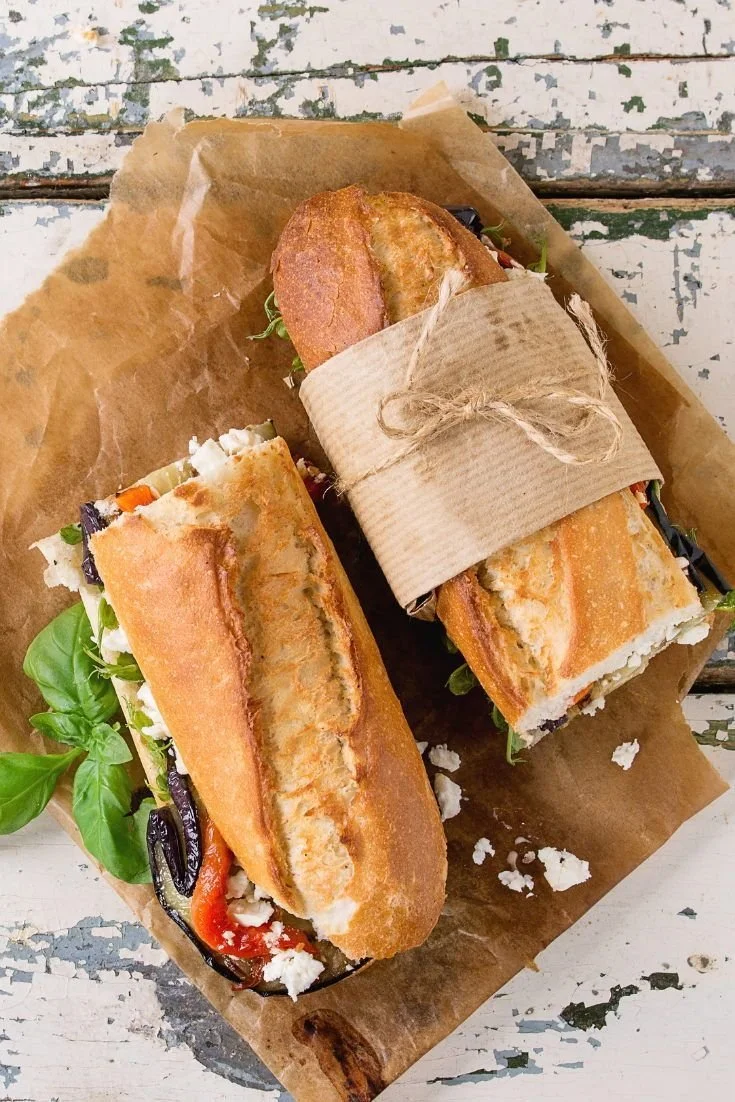 Two sandwich baguettes on brown parchment paper on a distressed white wooden surface, with one wrapped in brown paper and tied with twine. The sandwiches contain grilled vegetables and cheese.