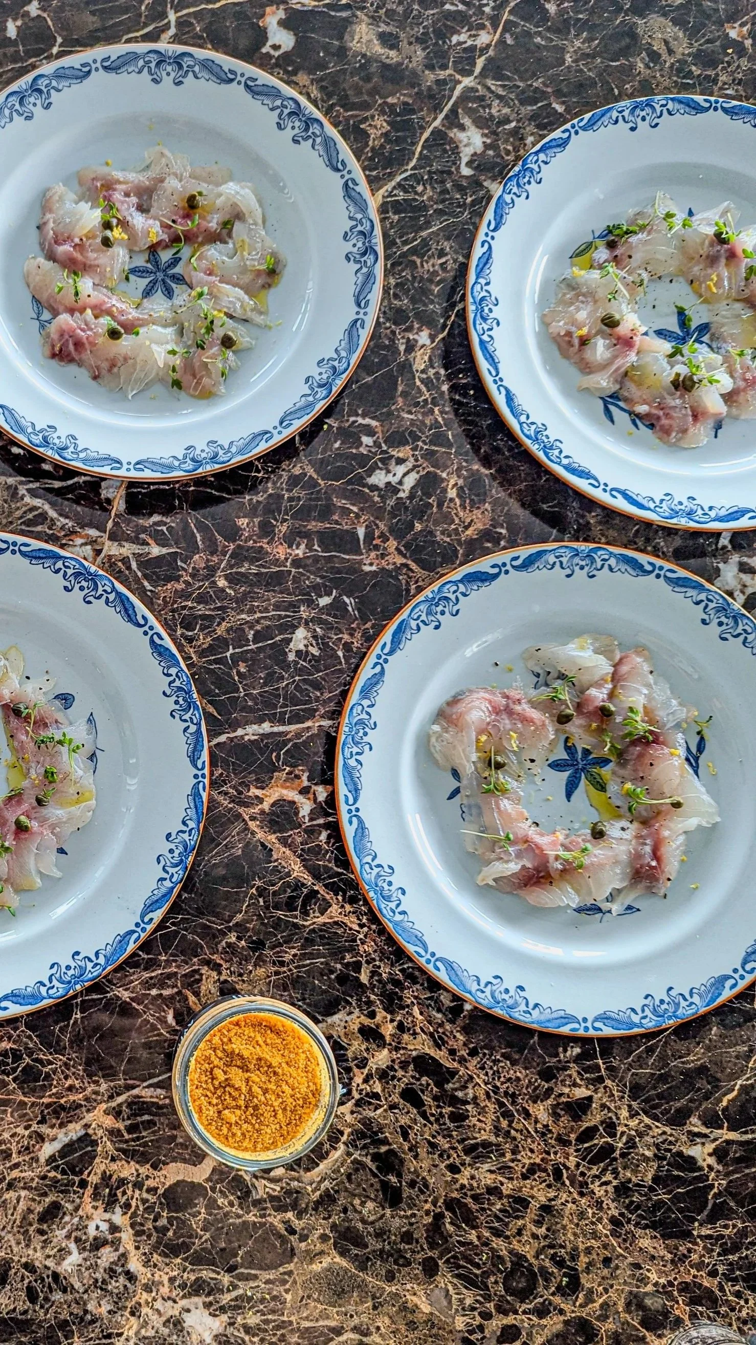 Four blue and white patterned plates with raw fish carpaccio garnished with herbs and capers, arranged on a dark marble table, with a small side of botarga to be served on the plates.