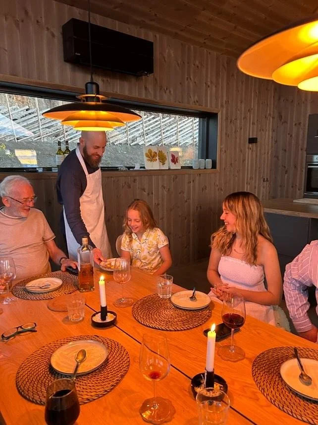 Family gathered around a wooden dining table enjoying a private dinner and specifically our dessert course with candles and wine glasses, celebrating while our private chef serves one of the guests his dessert plate.