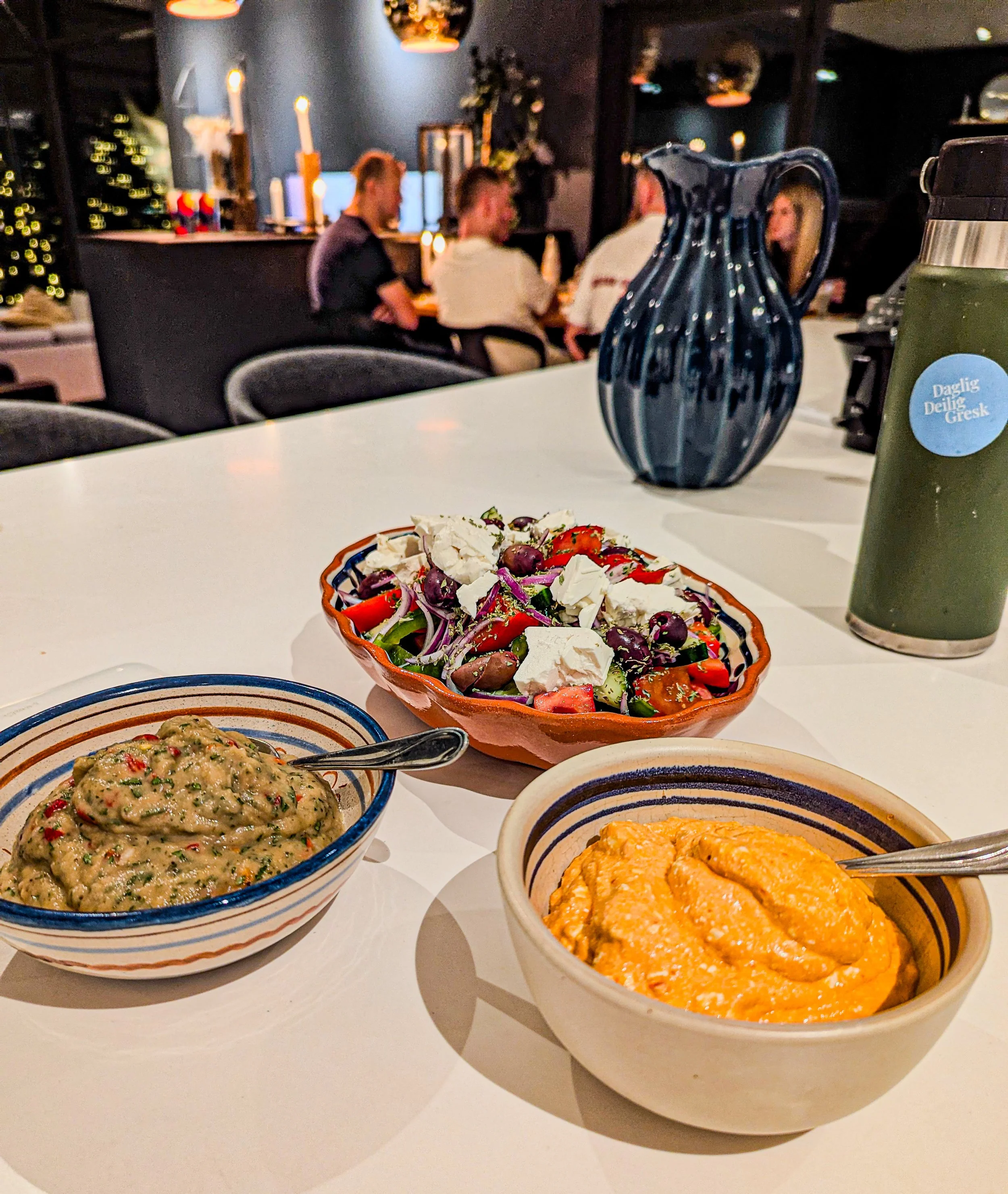 A table with three plates with starters, including a  salad with feta, a bowl of smoked eggplant dip, and a bowl of spicy feta dip. In the background our guests dining in a cozy, dimly-lit dining area enjoying our private dinner.