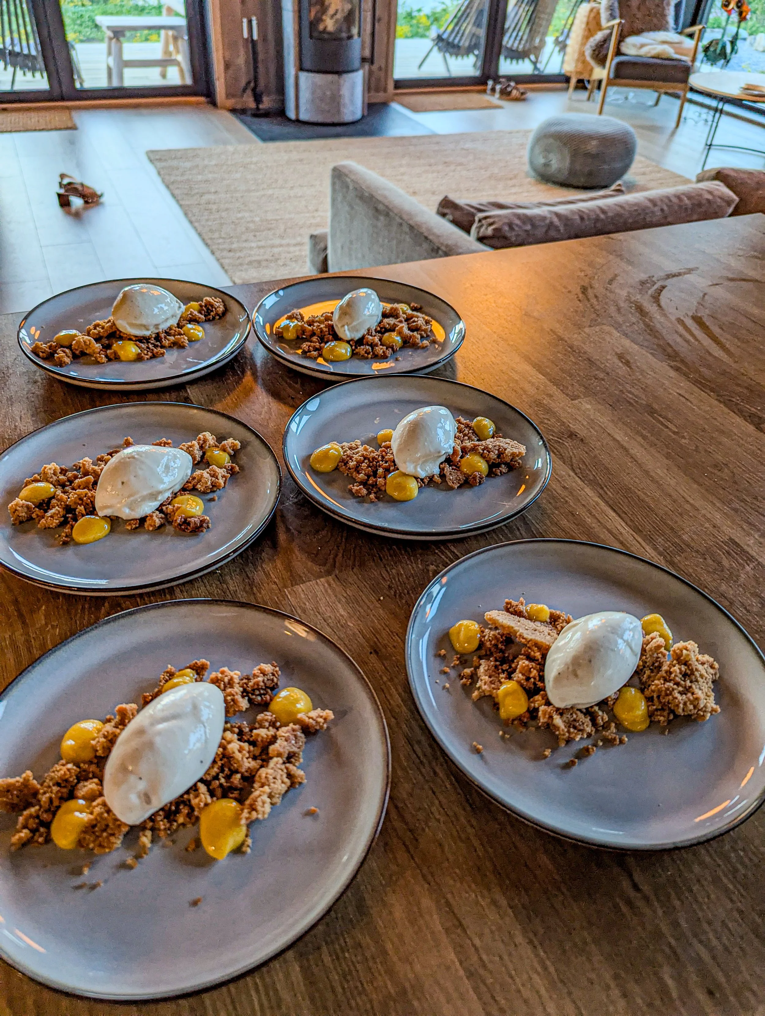Six dessert plates with a macadamia crumble, a scoop of vanilla ice cream, and lemon curd served on a wooden table in a cozy living room.