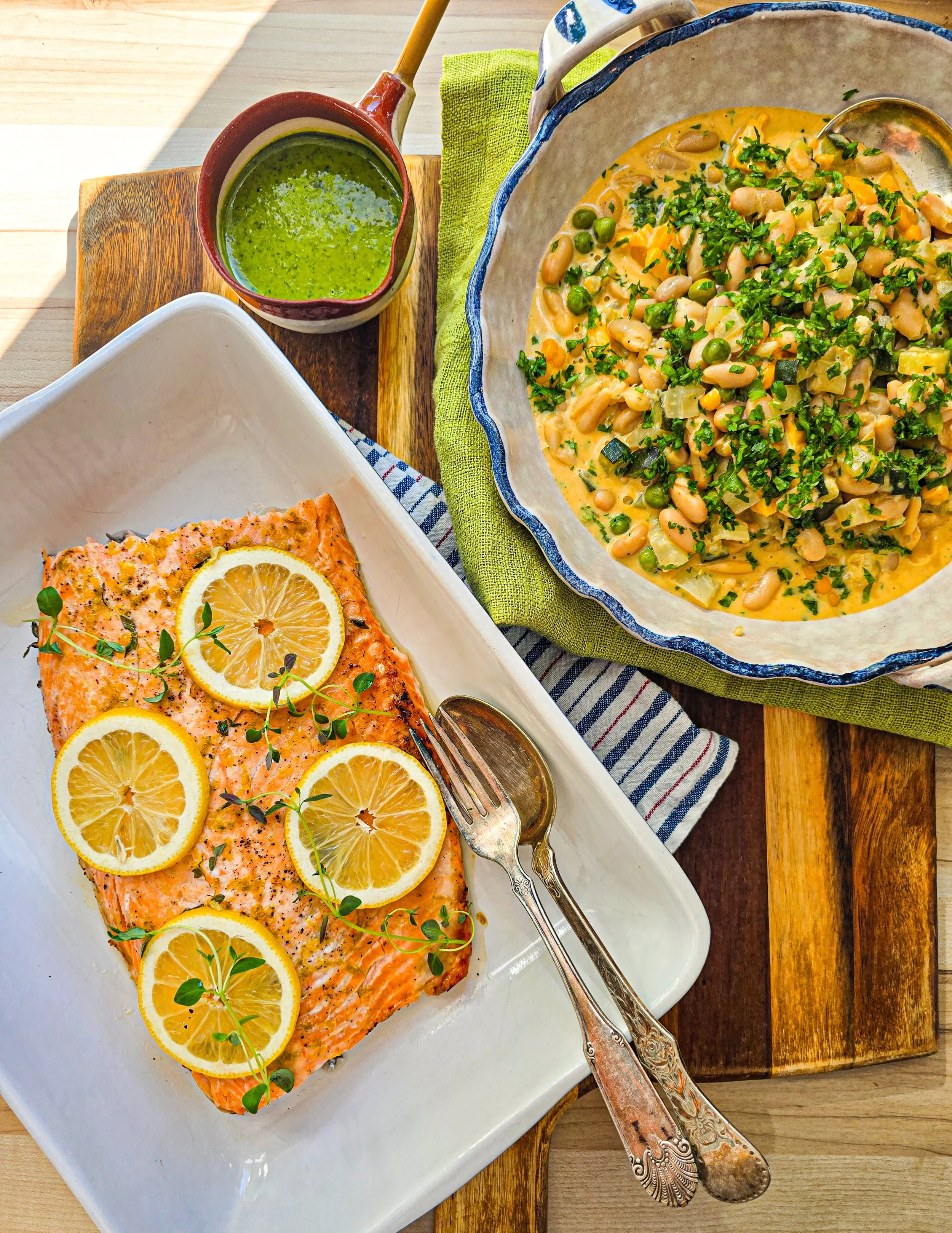 A plated baked salmon fillet garnished with lemon slices and microgreens, served with a side dish of beans cassoulet with spring vegetables and parsley, and a small pitcher with gremolata sauce, all arranged on a wooden table.