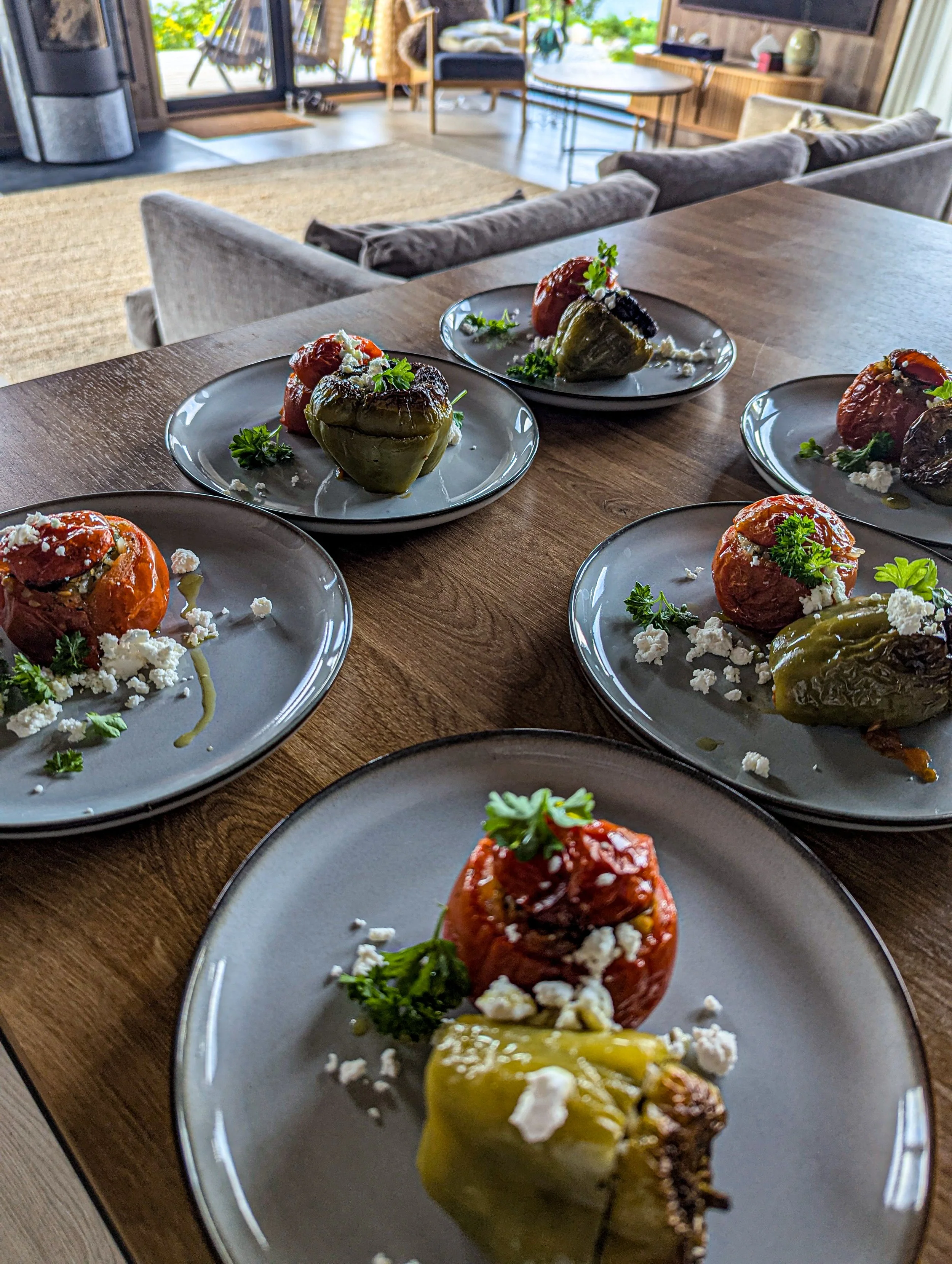 Seven plates of "gemista"-stuffed peppers & tomatoes garnished with parsley and crumbled feta on a wooden dining table in a bright, modern room with a cozy seating area and outdoor view.