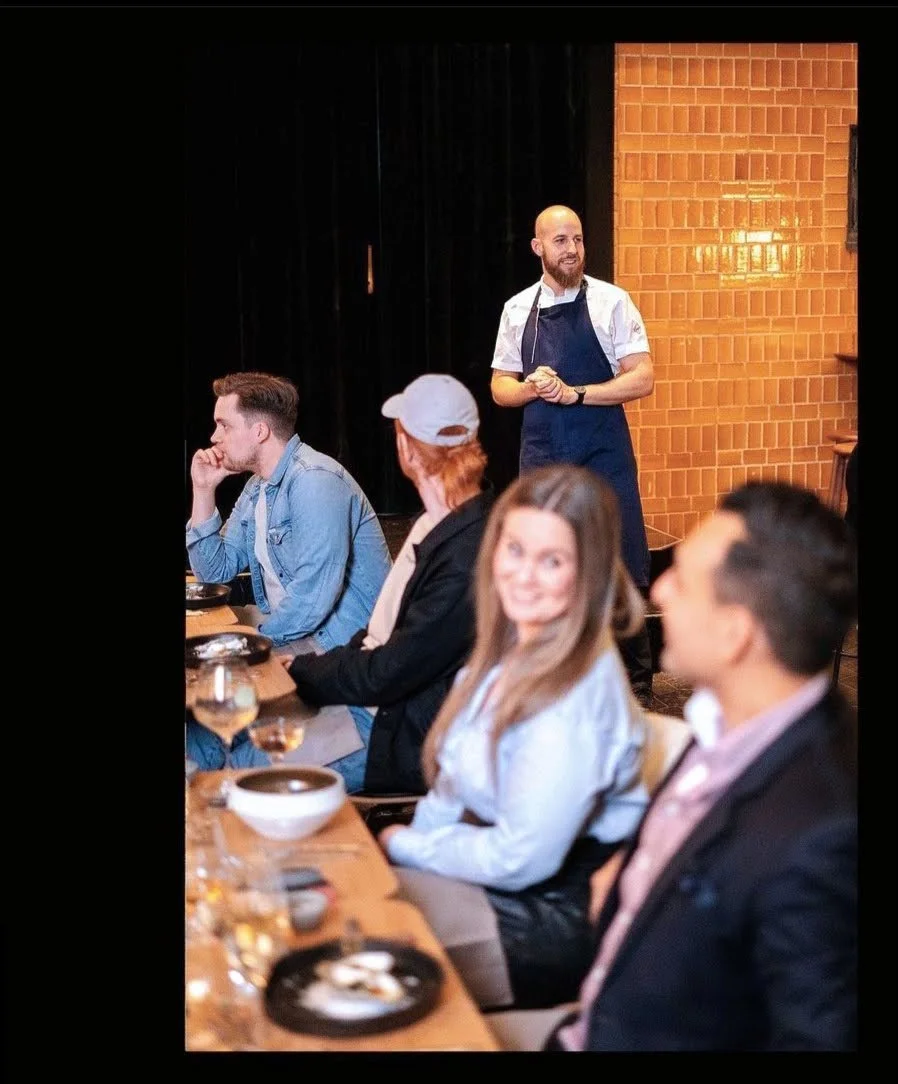 A private chef from noêma standing and explaing our nights menu to the  group of our guests seated at a dining table while enjoying their private dinner with us