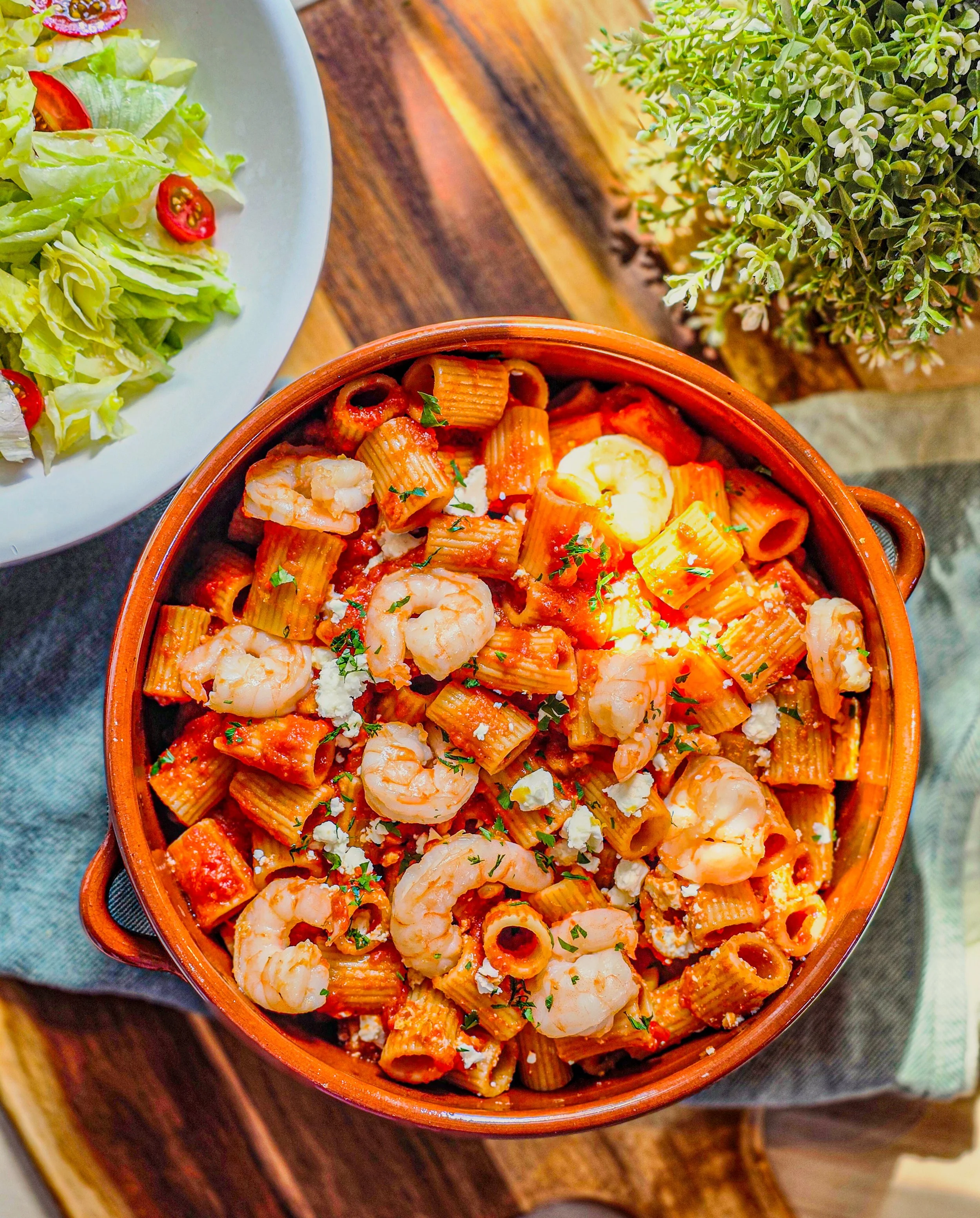 A terracotta bowl filled with rigattoni pasta with shrimps, and crumbled feta, garnished with herbs, on a wooden table. A white plate with a salad featuring lettuce and cherry tomatoes is served on the side.