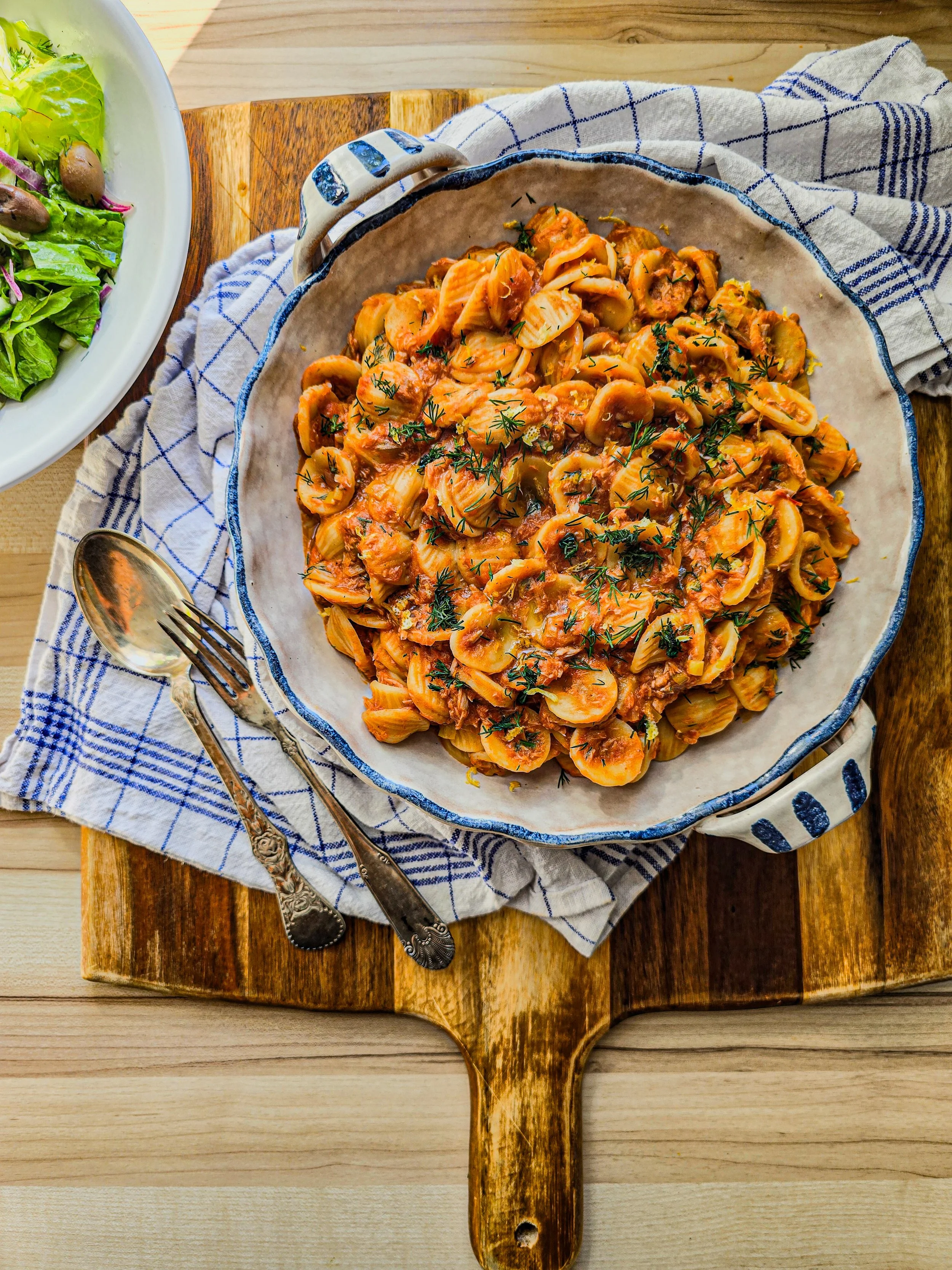 A bowl of casarecce pasta with tuna ragu with tomato based sauce and herbs on a wooden surface, with a salad nearby.