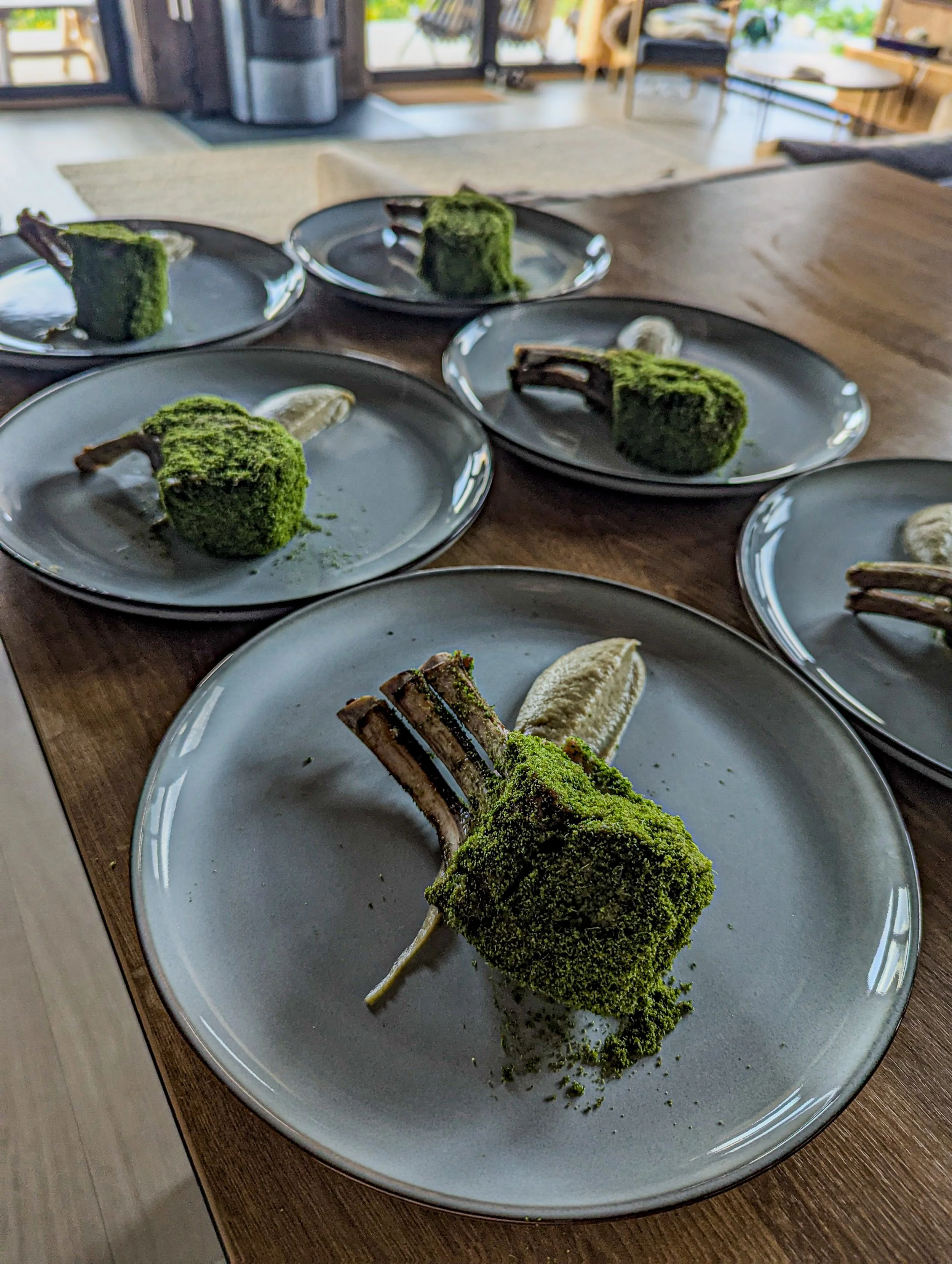 Six plates each with a roasted lamb chop coated in green herb crust and a dollop of smoked eggplant pure, arranged on a wooden dining table.