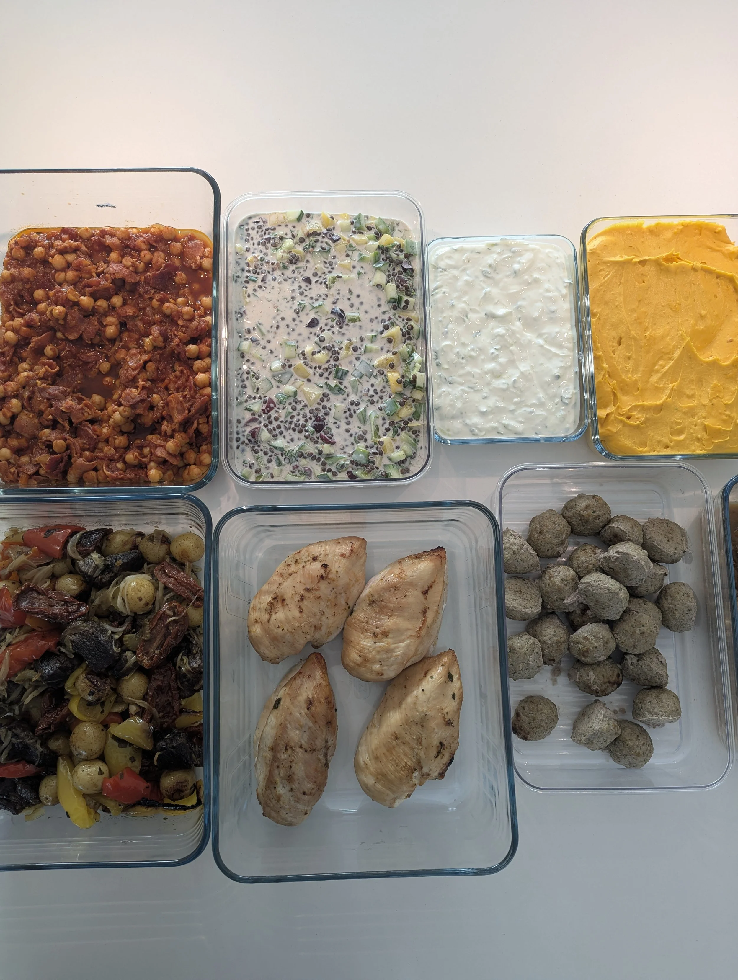A variety of food in glass containers from our weekly meal prep service, including chili, israeli lentils salad, tzatziki, pumpkin pure, roasted vegetables and italian meatballs, and grilled chicken breasts.