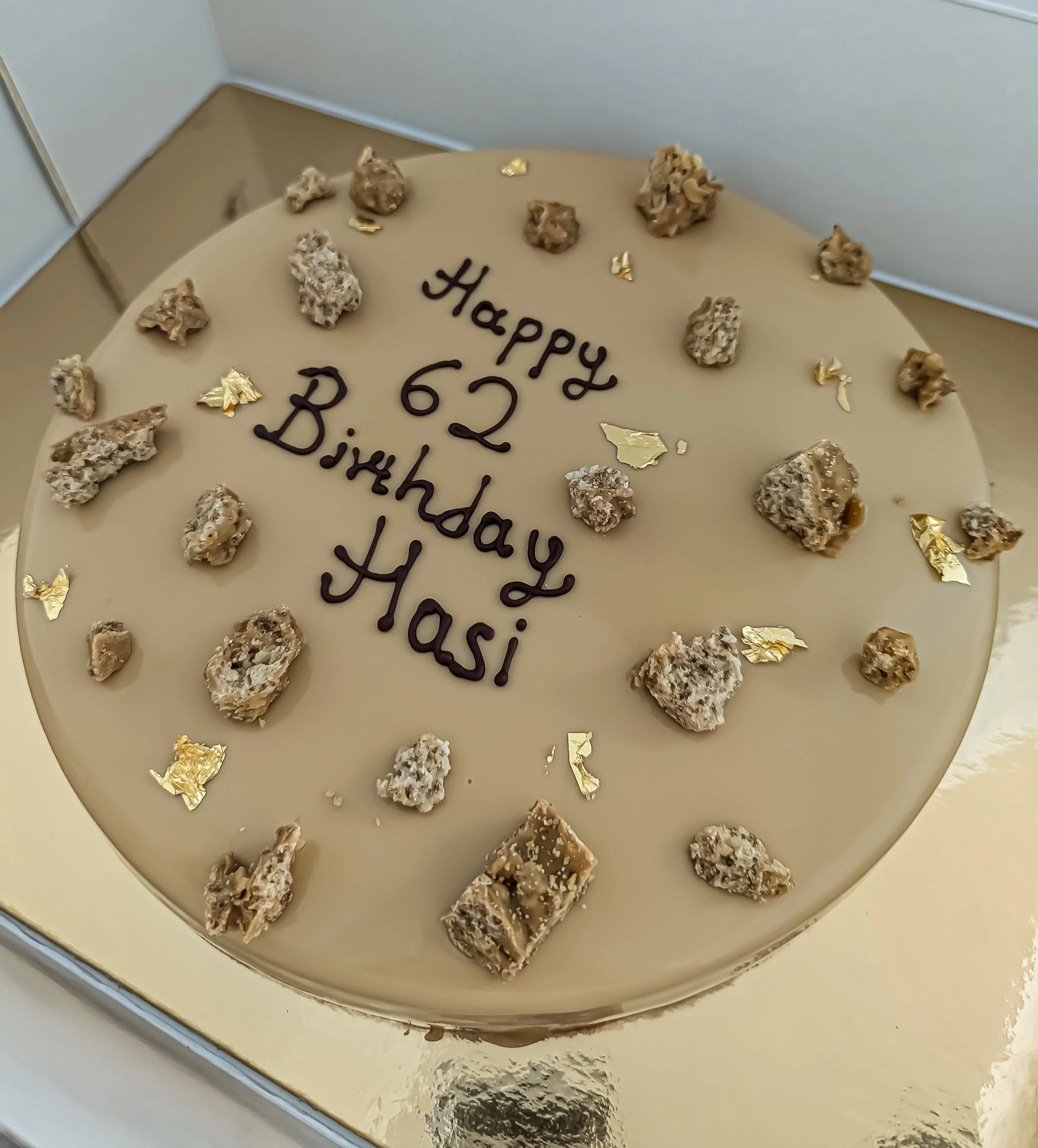 A round birthday cake with cream-colored icing, decorated with chocolate rocks and gold leaf pieces. The cake has a message written in chocolate that says, 'Happy 62nd Birthday Hasi'.