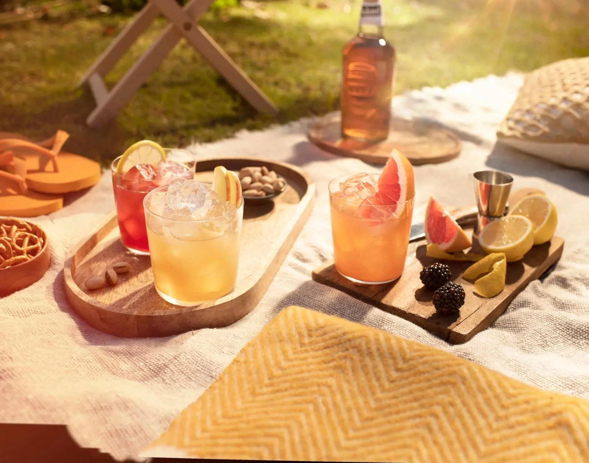 Outdoor picnic setup on a white tablecloth with colorful cocktails garnished with citrus slices and blackberries, a bottle of rosé wine, and various snacks and fruits in a sunny garden.