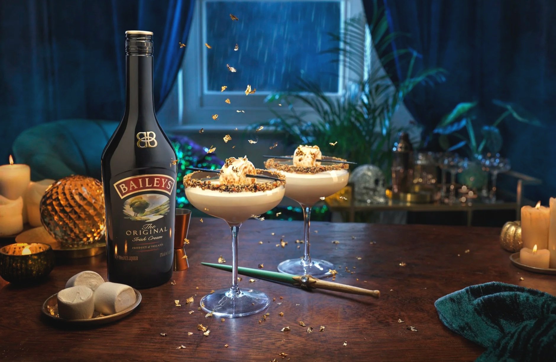 A bottle of Baileys Irish Cream liqueur, two dessert cocktails with whipped cream and sprinkles, marshmallows, candles, and teal slippers on a wooden table. A window with rain outside and dark blue curtains in the background.