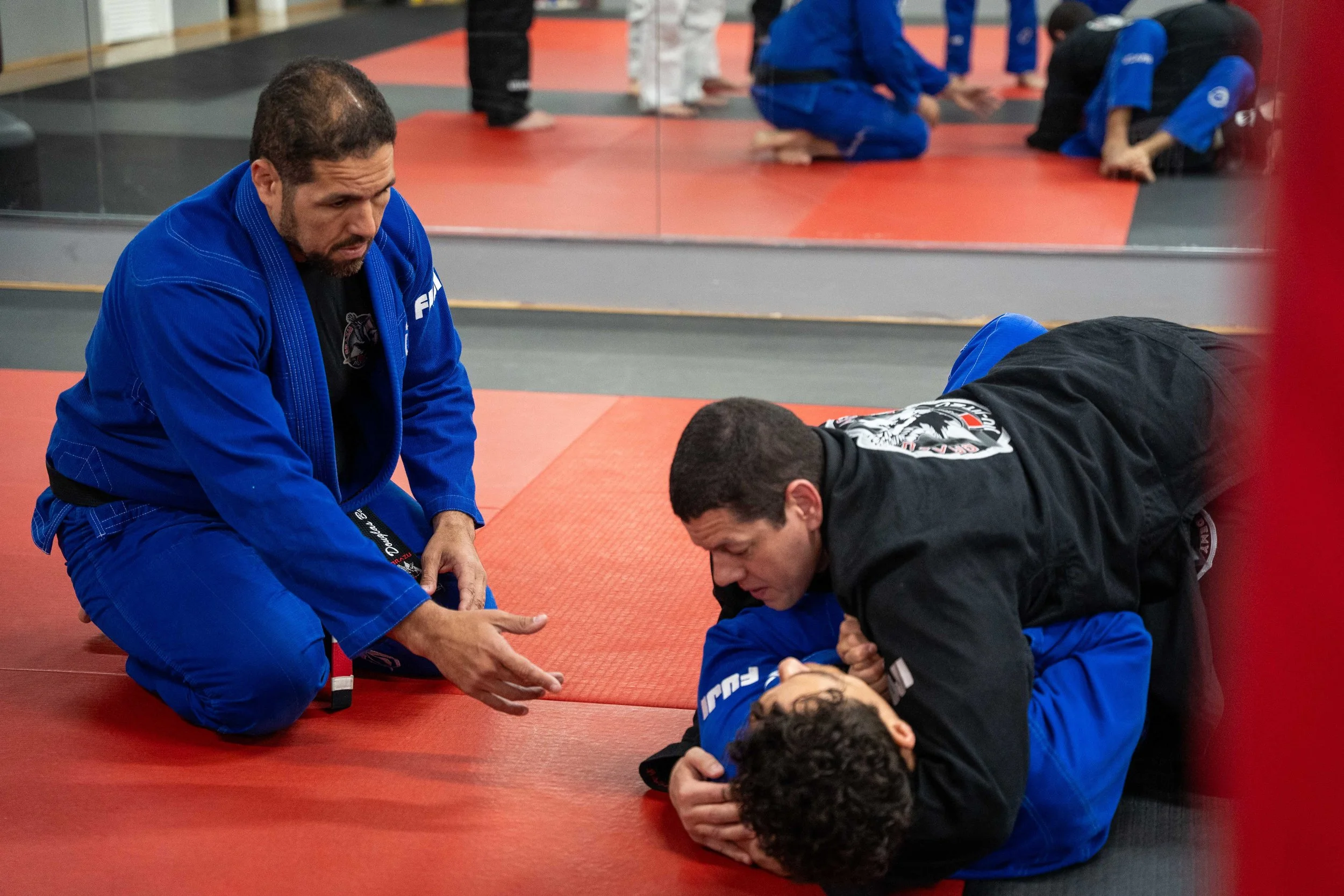 Brazilian Jiu-Jitsu practitioners practicing on red mats, with a coach instructing a student in a black gi, and other students training in the background.