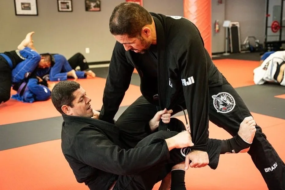 Two men practicing Brazilian Jiu-Jitsu on a mat, with other practitioners training in the background.