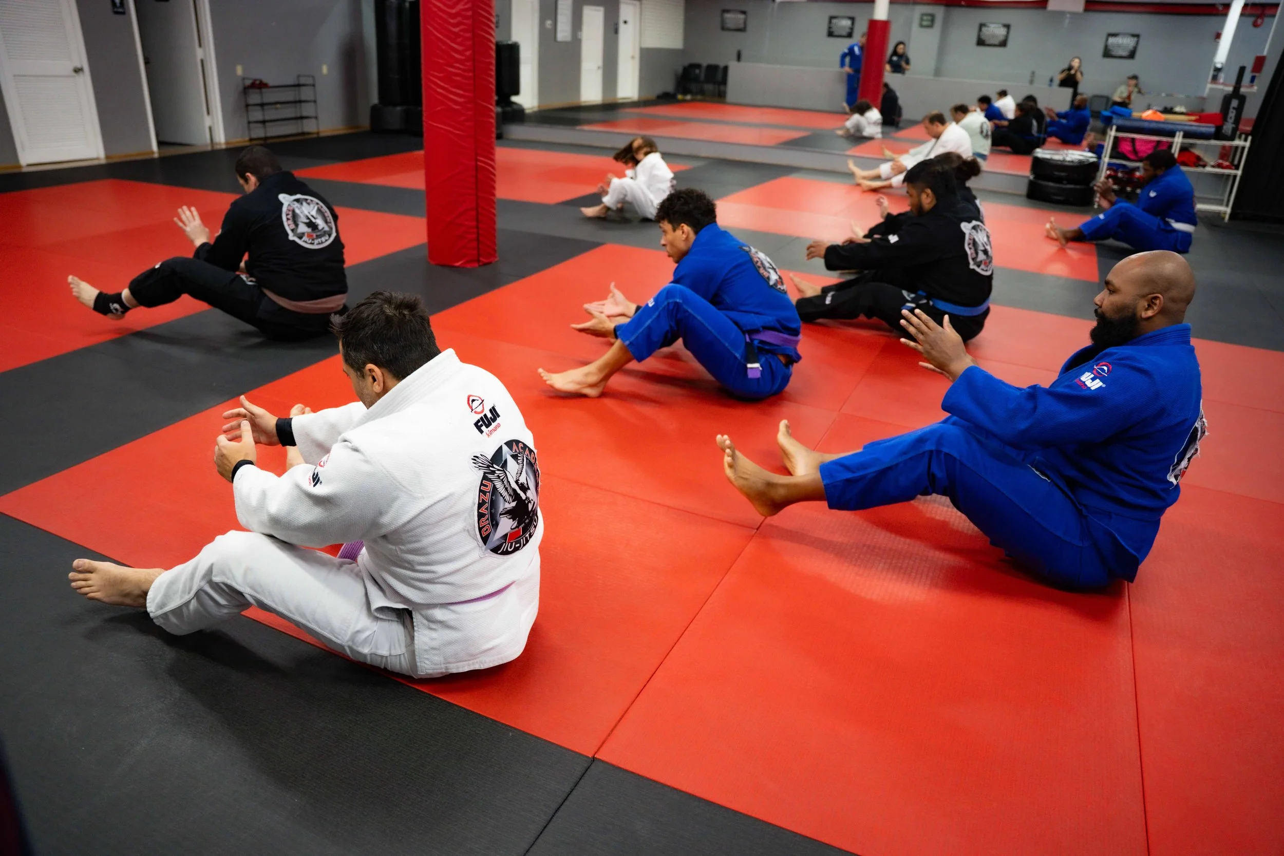 Group of people practicing Brazilian Jiu-Jitsu on red and black mats in a martial arts gym, wearing traditional gi uniforms of various colors.