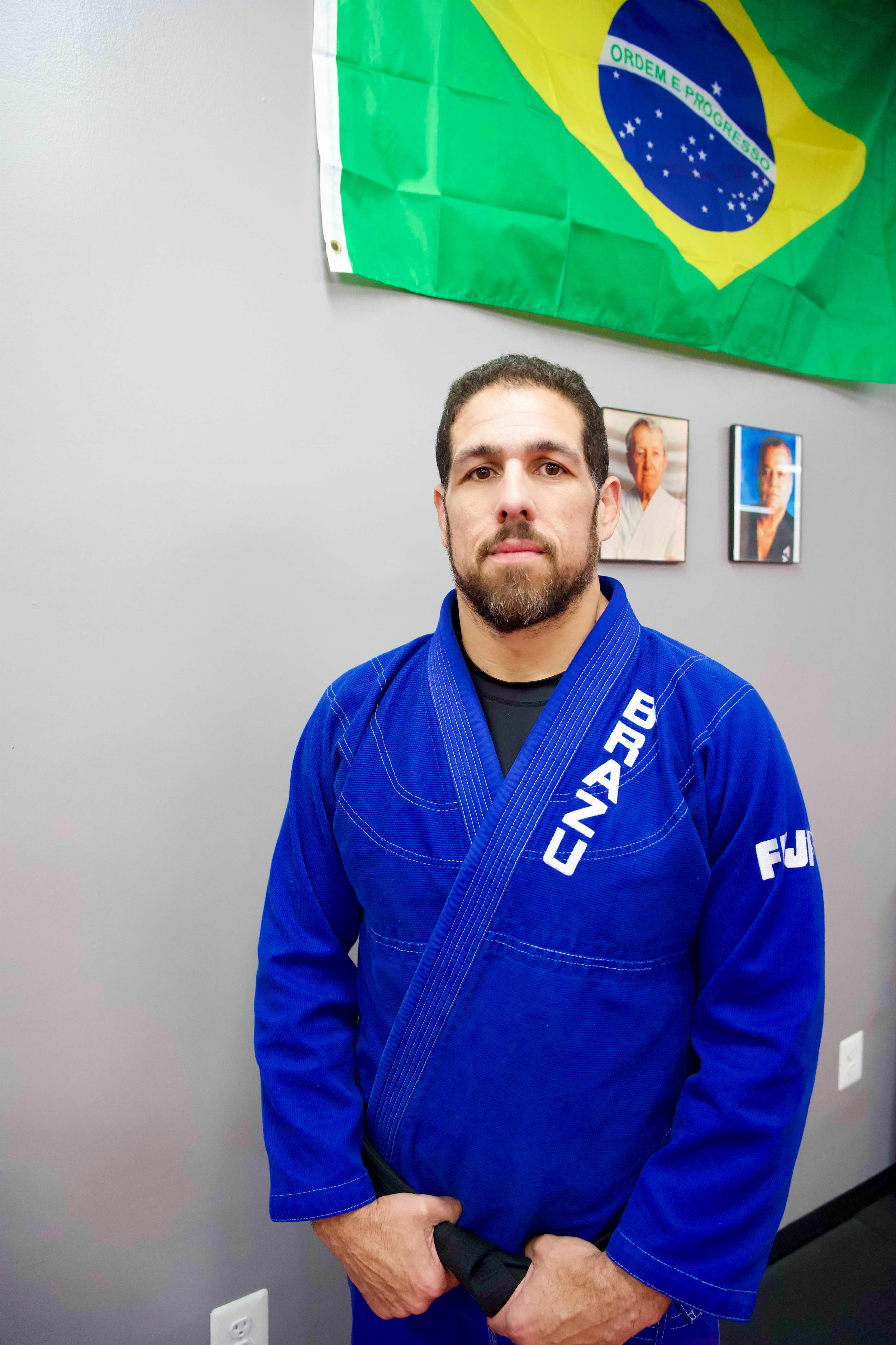 Black belt Douglas Barros wearing a blue  Brazu Brazilian Jiu-Jitsu gi standing in front of a gray wall with a Brazilian flag and framed pictures.