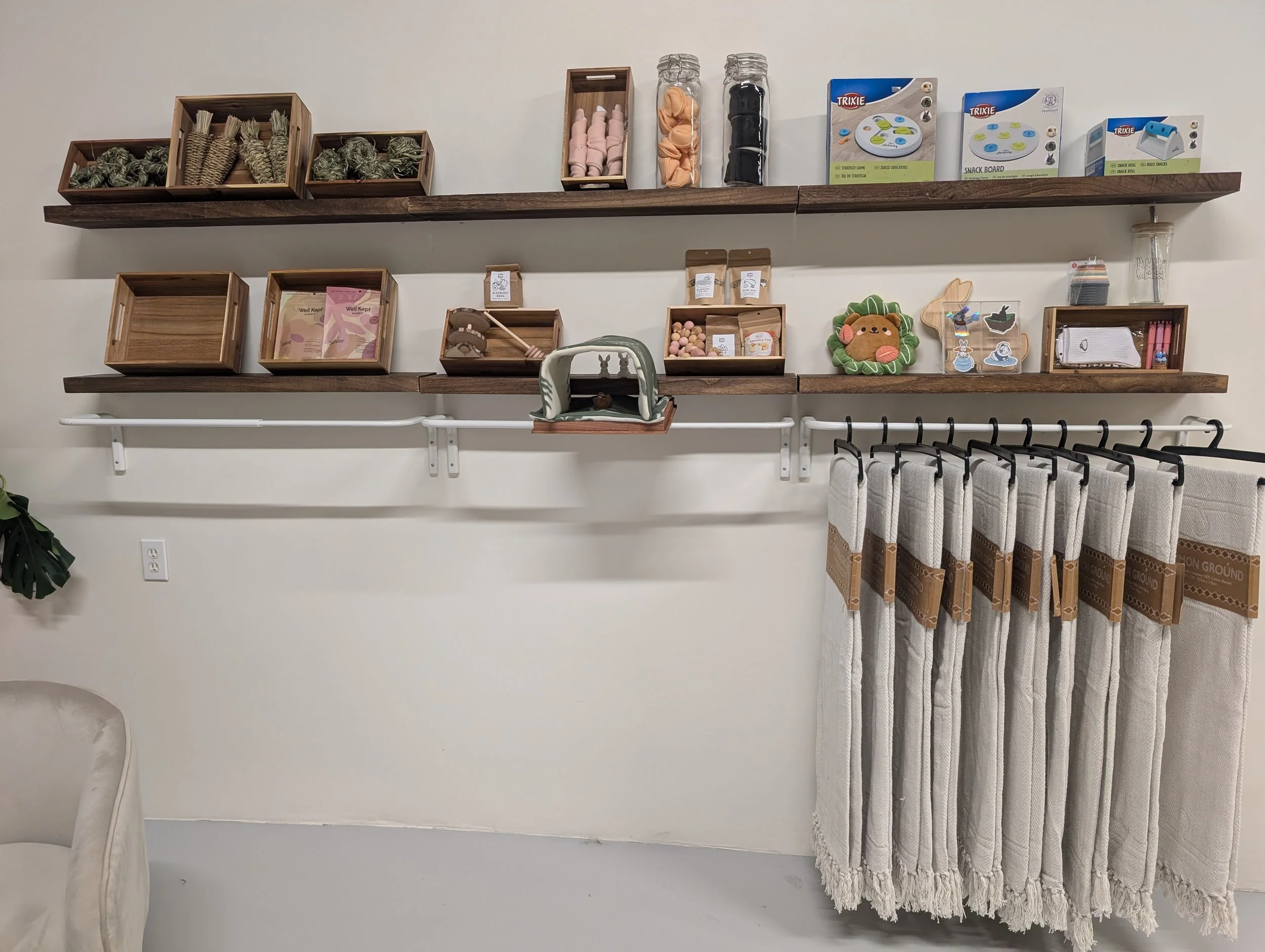The Burrow Exchange: Wooden shelves with various items including snacks, toys, and decor on a white wall, with a white curtain hanging below and a partial chair visible on the left.