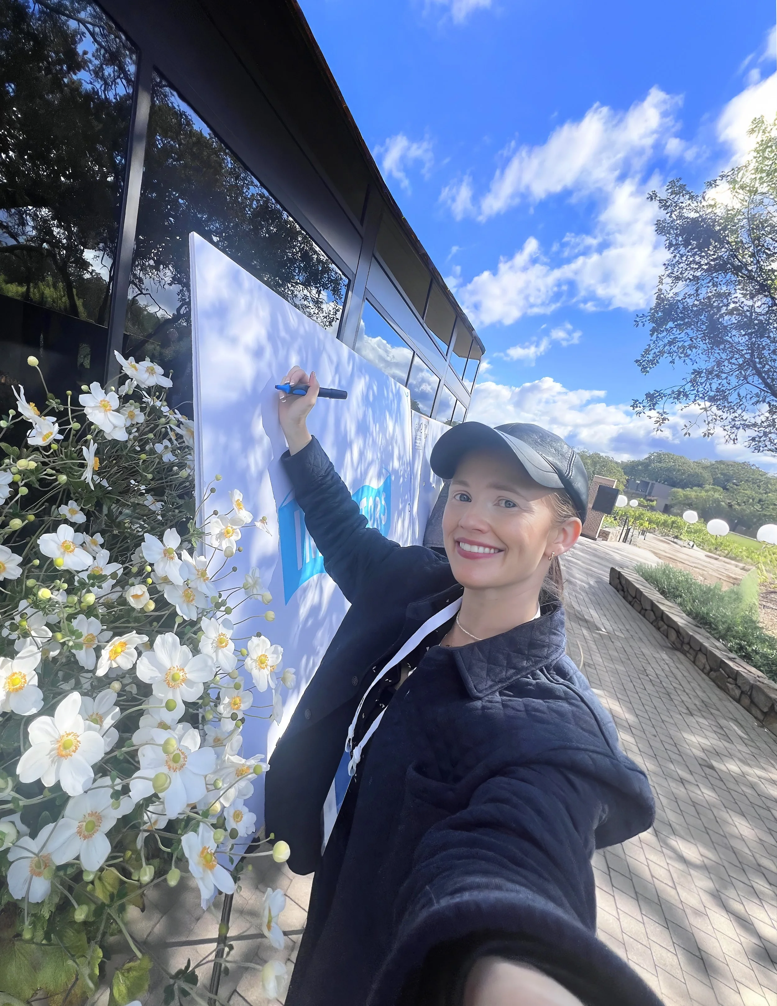 Smiling woman in black jacket and cap signing a large outdoor sign with a marker near white flowers and a glass building on a sunny day.
